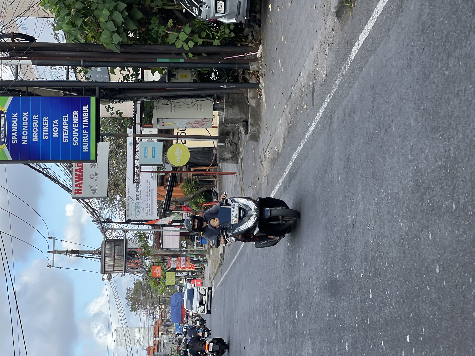 The image captures a street scene in Denpasar, Indonesia, likely on a main road given the traffic and signage. In the foreground, a person is riding a black scooter, with a young child seated in front of them. Both individuals are wearing face masks and helmets for protection. The scooter has a license plate displaying "DK 5716 AED."
The street is paved and appears to be moderately busy with other vehicles, including a white car and several other scooters, in the background. Overhead, a complex network of electrical wires and utility poles dominates the sky, a common sight in urban Indonesian environments.
Along the side of the road, various signs are visible, advertising services such as "HAWAII," "YAMAHA," and "SPANDUK NEONBOX BROSR STIKER NOTA STEMPEL SOUVENER HURUF TIMBUL." These indicate a commercial area with businesses offering printing, signage, and potentially other related services. A silver car is parked on the right side of the road. The overall environment suggests a typical bustling street in a developed city in Indonesia.