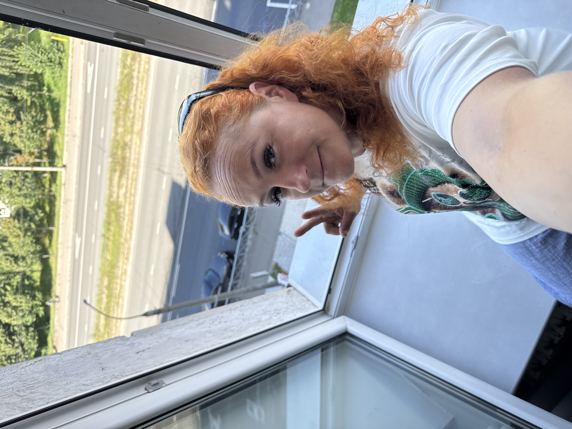 A woman with ginger hair is taking a selfie from a balcony, with a road visible in the background. She is wearing a white t-shirt and blue jeans, and has sunglasses on her head. Her left hand is raised, making a peace sign. In the background, there are trees, cars, and a street lamp. The image appears to have been taken in the city of Kazan, Russia, based on the architecture and vegetation visible.