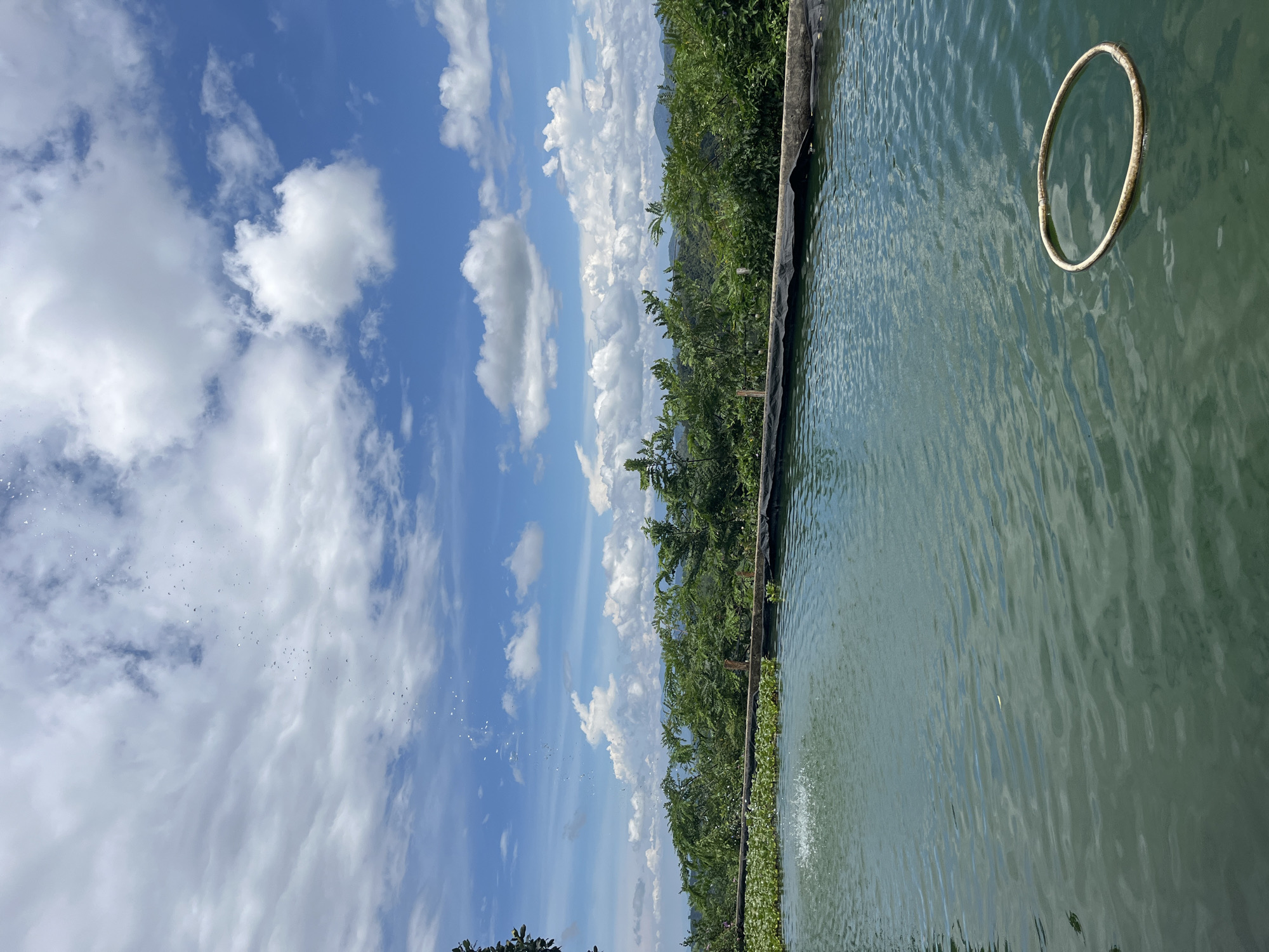 A high-angle, full shot captures an outdoor pond with a hazy, distant view of lush green hills under a bright blue sky dotted with white, puffy clouds. In the foreground, the water is a tranquil, murky green, with gentle ripples creating a pattern on its surface. To the left, a small fountain sprays water upwards, creating a misty arc that reaches towards the sky. A circular, floating ring with a light brown, textured surface rests on the water in the lower right quadrant. A low, dark, rough-edged wooden border runs along the edge of the pond, separating it from a dense line of green foliage and trees. The foliage forms a verdant wall, behind which layers of hills recede into the distance, softened by a light atmospheric haze. The overall scene evokes a sense of calm and natural beauty, suggesting a peaceful retreat in Sơn Điền, Vietnam.