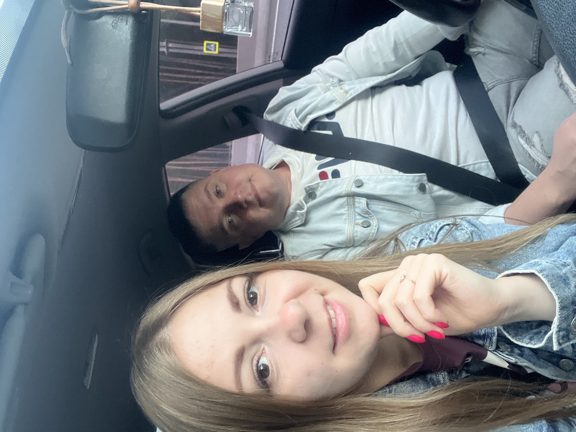 The image shows two individuals, a woman and a man, in the front seats of a car. The woman is in the foreground, smiling at the camera, with her hand near her chin, adorned with bright pink nail polish and a ring. She is wearing a denim jacket over a dark shirt. The man is to her right, also looking towards the camera, wearing a light-colored denim jacket over a white t-shirt with lettering. Both individuals appear to be wearing seatbelts. The interior of the car is visible, including the dashboard and a rearview mirror. Outside the car, through the windows, a blurred background suggests an urban environment, with some vertical elements that could be trees or buildings. A car air freshener is hanging from the rearview mirror. The presence of a yellow pedestrian crossing sign outside the car window might indicate a location within a city. There is no specific information in the image to determine the location as Allaki, Russia.