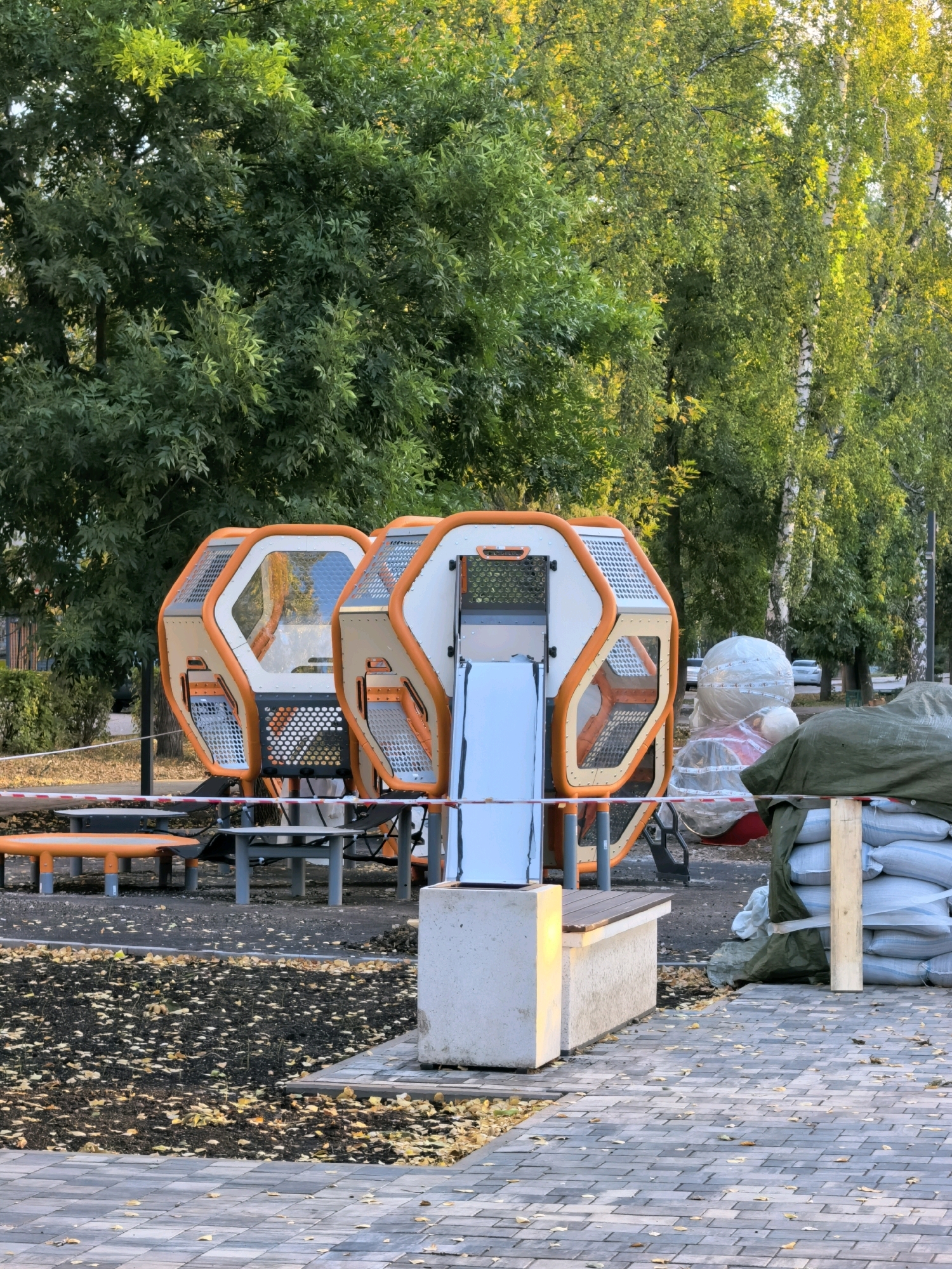 The image depicts an outdoor scene in what appears to be a park or public area, likely in Sterlitamak, Russia, given the context provided. In the foreground, a paved walkway made of grey and tan interlocking bricks is visible, scattered with fallen yellow leaves. To the left, a patch of dark soil is also covered with leaves, bordering a light-colored concrete structure with a wooden bench.
Dominating the mid-ground is a modern playground structure with two interconnected hexagonal pods, featuring white panels, orange frames, and transparent sections. A light blue slide extends from one of the pods. Red and white striped caution tape is strung across the area in front of the playground equipment. To the right of the playground, several large white sacks are stacked and covered with a dark green tarp, supported by wooden beams, suggesting ongoing construction or flood control measures. A cylindrical object wrapped in transparent plastic is also visible behind the sacks.
The background consists of lush green trees, with some branches showing yellowing leaves, indicating it is likely autumn. The overall impression is a public space in transition, with elements of play and temporary protective measures present. No people are visible in the image.