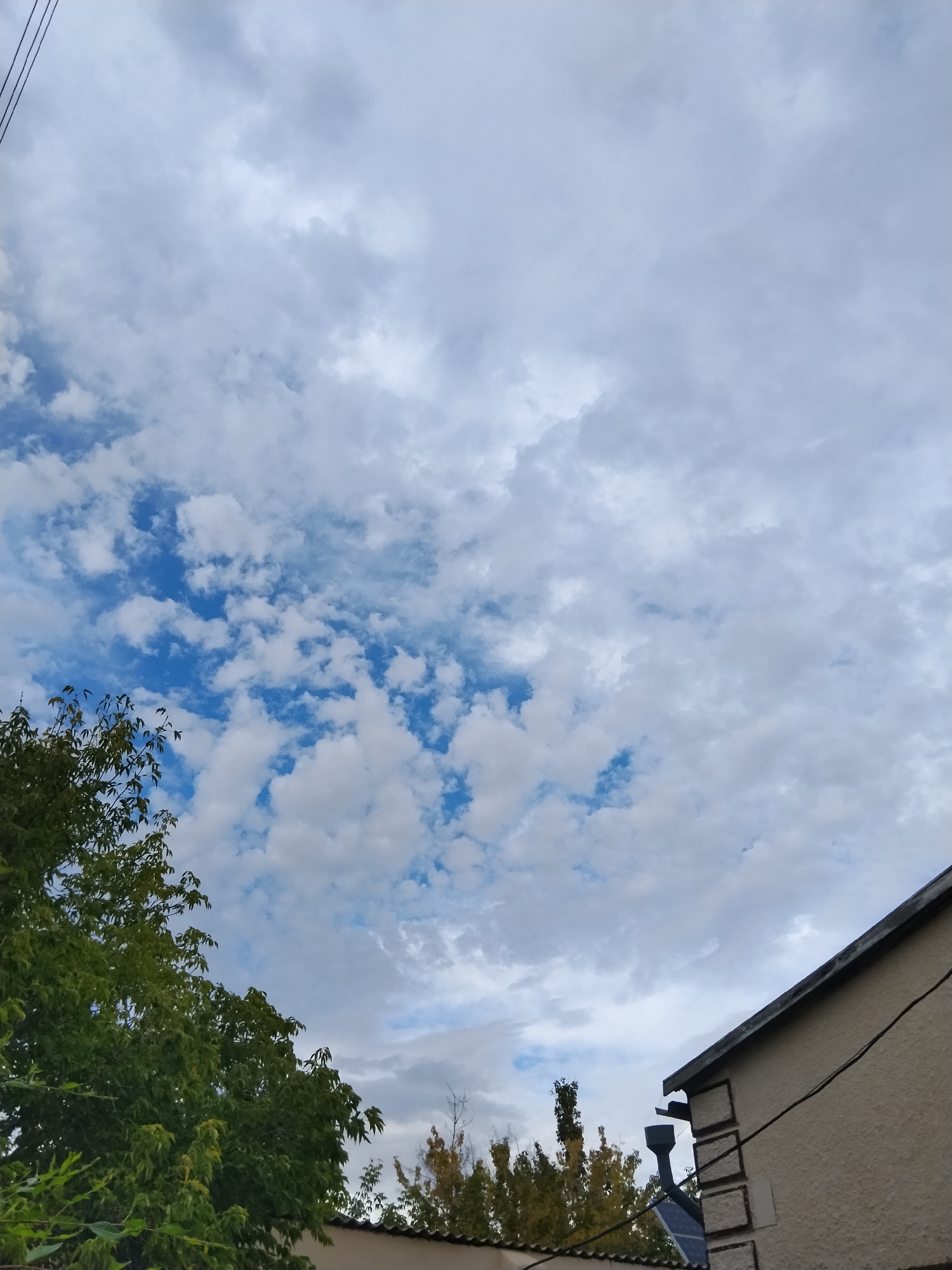 The image depicts an outdoor scene with a cloudy sky dominating the frame. In the foreground, a portion of a building's roof and wall is visible, featuring a dark-colored gutter system and what appears to be a solar panel attached to the wall. To the left, the lush green foliage of trees fills the lower left quadrant, suggesting the presence of vegetation. The sky itself is a mix of white and gray clouds, with patches of blue sky peeking through. The overall impression is that of a slightly overcast day. There are no people or animals visible in the image, nor any specific identifiable landmarks that would allow for pinpointing a location within Kokshetau, Kazakhstan. The environment appears to be residential, given the presence of a building and trees.