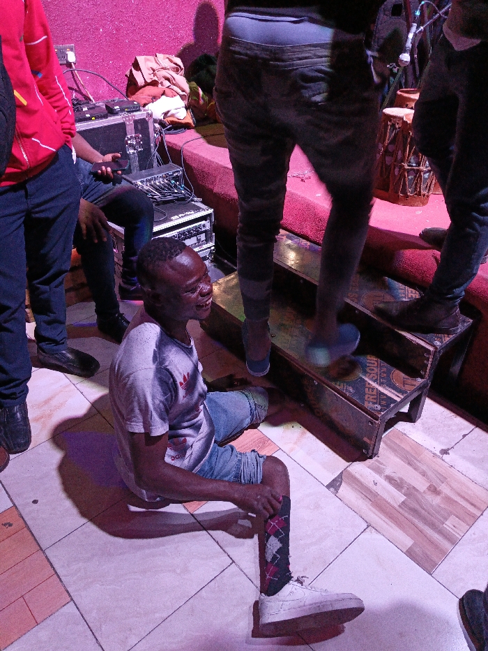 In a candid shot bathed in violet lighting, a man in a white t-shirt and denim shorts sits on a tiled floor, one leg outstretched in an argyle sock and white sneaker. He's smiling, partially obscured by shadows. Above him, two figures stand on makeshift wooden steps leading to a red-carpeted platform. Other individuals flank him, including one in a red hoodie and another in dark pants holding a phone, all near sound equipment cases.