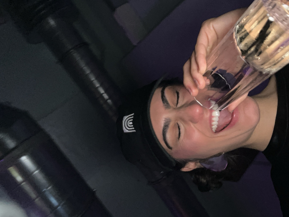 A person wearing a black beanie and a black shirt holds a transparent glass up to their mouth, as if about to drink from it. Their eyes are closed and their mouth is open, with teeth visible. The background consists of a dark, textured surface, possibly a wall or a pipe system. The overall image has a slightly tilted perspective.