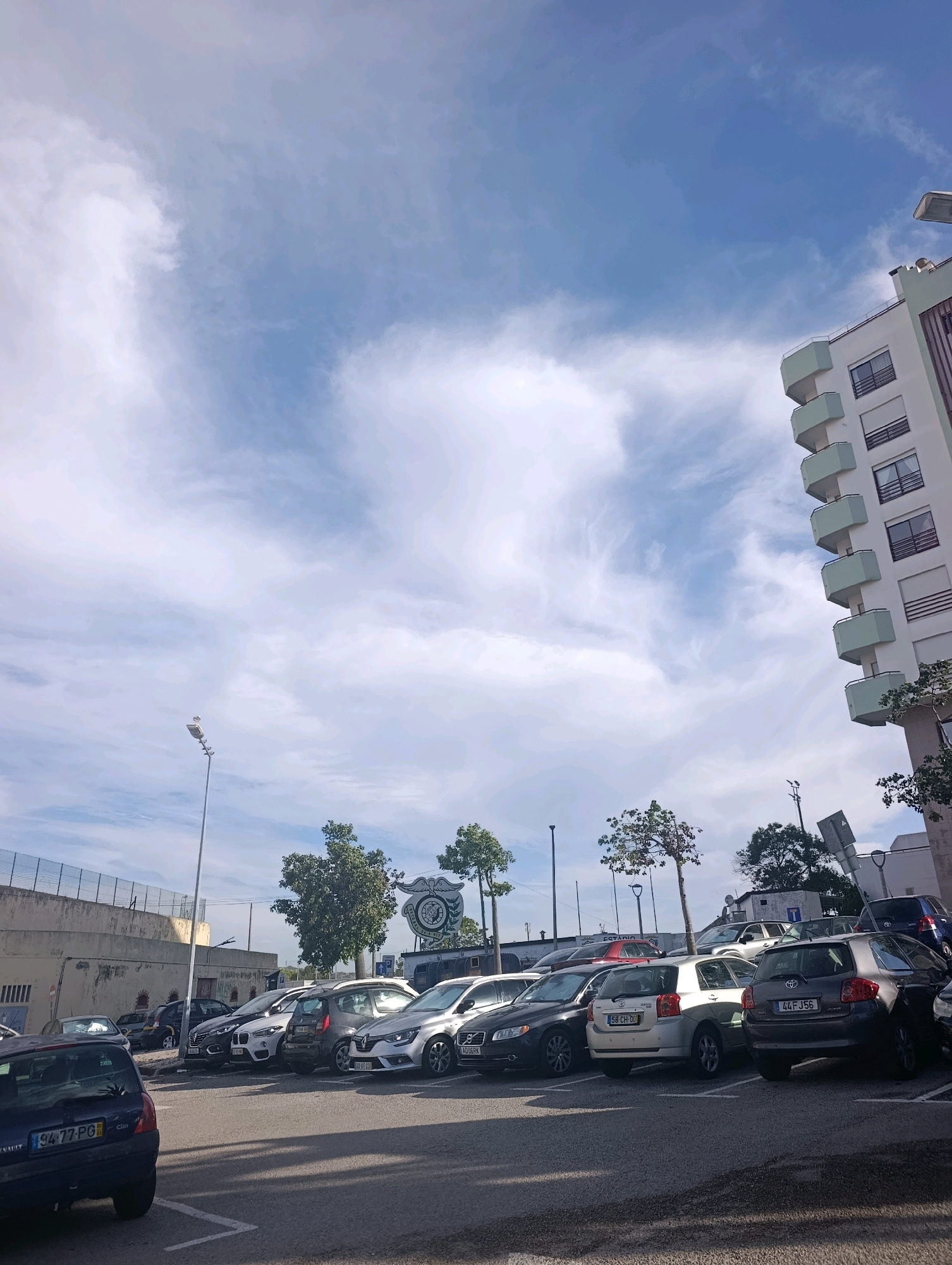 A medium shot captures a parking lot filled with various vehicles under a partly cloudy sky in Setúbal, Portugal. The scene is dominated by a row of parked cars, predominantly sedans and hatchbacks, some with visible license plates. Among them are a dark blue Renault Clio, a silver Renault Captur, and a silver Toyota Auris with license plate "44 FJ56". In the background, a modern building with light green balconies stands to the right, while a stadium fence borders the left side of the frame. Trees dot the landscape, and a street light stands tall. A large circular emblem with a logo is visible behind the cars, suggesting a sports venue or club. The time of day appears to be daytime, with sunlight casting shadows on the asphalt. No individuals are present in the image.