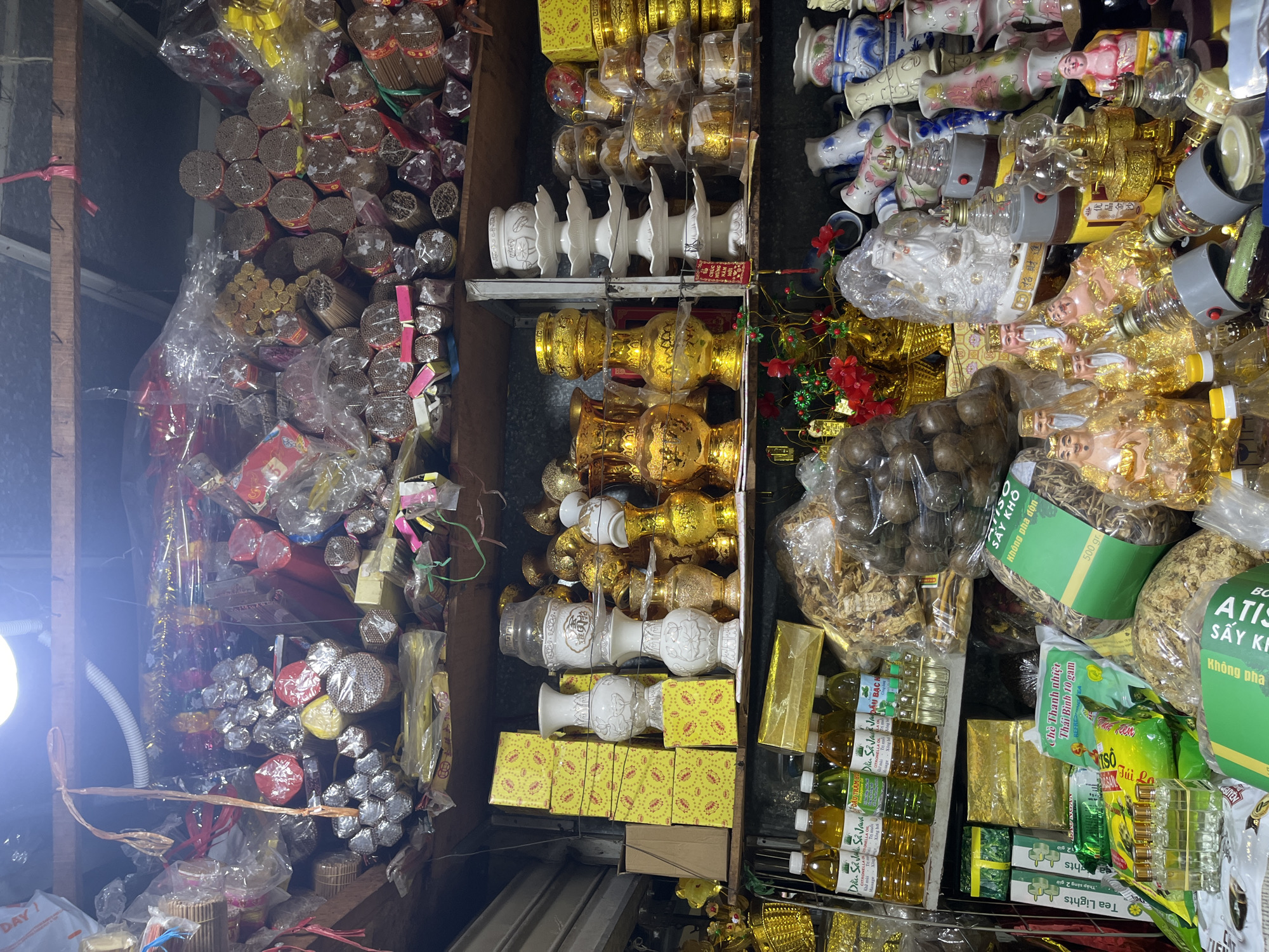 The image shows a cluttered stall in Nha Trang Ward, Vietnam, likely a shop selling religious or spiritual items. The stall is packed with various objects, including stacks of incense sticks, decorative jars and vases, golden figurines, and bottled oils. The lighting suggests an indoor setting, possibly a market or a small store. The overall impression is one of abundance and variety, with items filling multiple shelves from floor to ceiling.