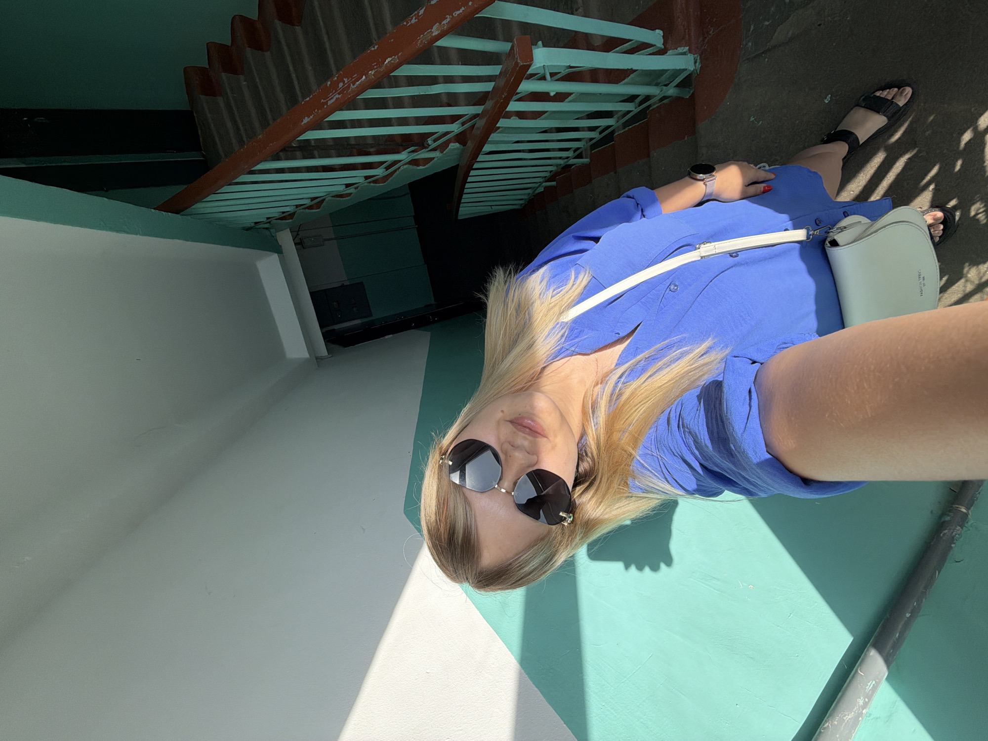 A downward, canted, selfie shot shows a woman in an environment featuring an architectural feature. The woman is fair-skinned, has blonde hair and is wearing sunglasses and a blue dress with a white shoulder bag. Her feet are clad in black sandals. She wears a wristwatch on her left wrist and has red nail polish.
The architectural feature is a set of stairs with reddish-brown supports and light blue slats that has a doorway underneath it. The woman is standing on a platform with white and light blue diagonal sections. The environment appears to be outdoors in daylight. The city of Chaikovsky, Russia is not visible, but is provided as the implied location.