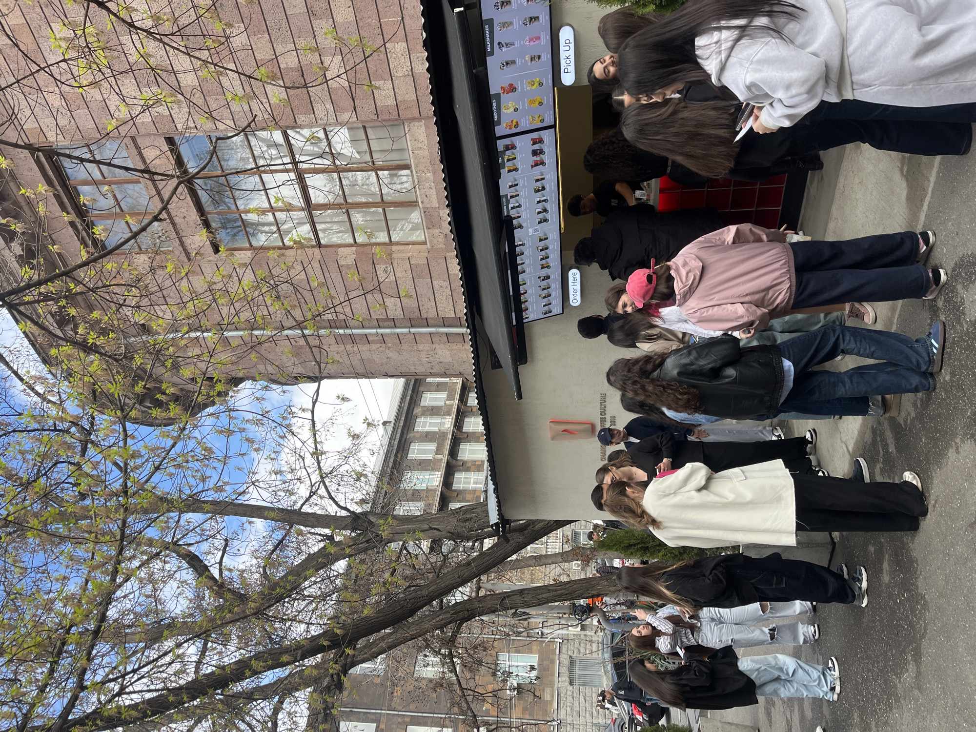 The image shows a long line of people queuing outside a shop in Yerevan, Armenia. The shop has a menu board displaying various drink options like smoothies and milkshakes. The line extends along the sidewalk, with individuals of varying ages and attire. The environment includes trees with sparse leaves, revealing a partly cloudy sky above. Buildings with multiple windows are visible in the background.