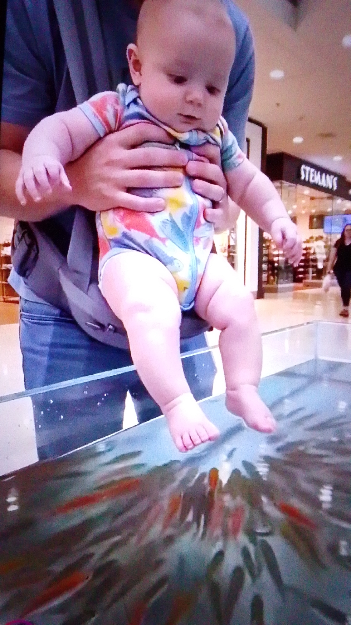 A baby is held by an adult, their feet dangling over a tank filled with small, colorful fish. The baby is wearing a brightly patterned onesie. The background suggests an indoor public space, possibly a mall, with storefronts visible. One storefront has a sign that reads "STEMANS". The lighting is bright, illuminating the scene and the water in the tank. The focus is on the baby and its interaction with the fish, which appear to be attracted to its feet. The overall impression is one of playful curiosity and a unique sensory experience. There is no information in the image to indicate the location is Aznakayevo, Russia.
