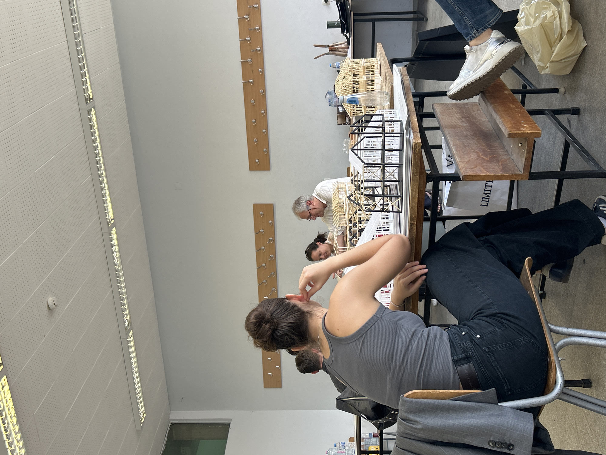 The image depicts an indoor setting, likely a classroom or workshop, with several individuals engaged at tables laden with architectural models. The room is brightly lit by fluorescent ceiling lights and features white walls with wooden coat racks. In the foreground, a woman with her hair in a bun is seated at a table, wearing a grey tank top and dark pants. She is looking towards her right, with her hand near her ear. On the table in front of her are various architectural models made of wood and black structural elements, along with bottles of water. To her right, another person, partially visible, is wearing white sneakers and jeans. Further into the scene, an older man with glasses and a white shirt is seated at a table, also surrounded by architectural models. Another woman is seated beside him, smiling. The overall atmosphere appears to be one of focused work or a class session. The image does not contain any explicit visual cues to pinpoint its location in Sofia, Bulgaria.
