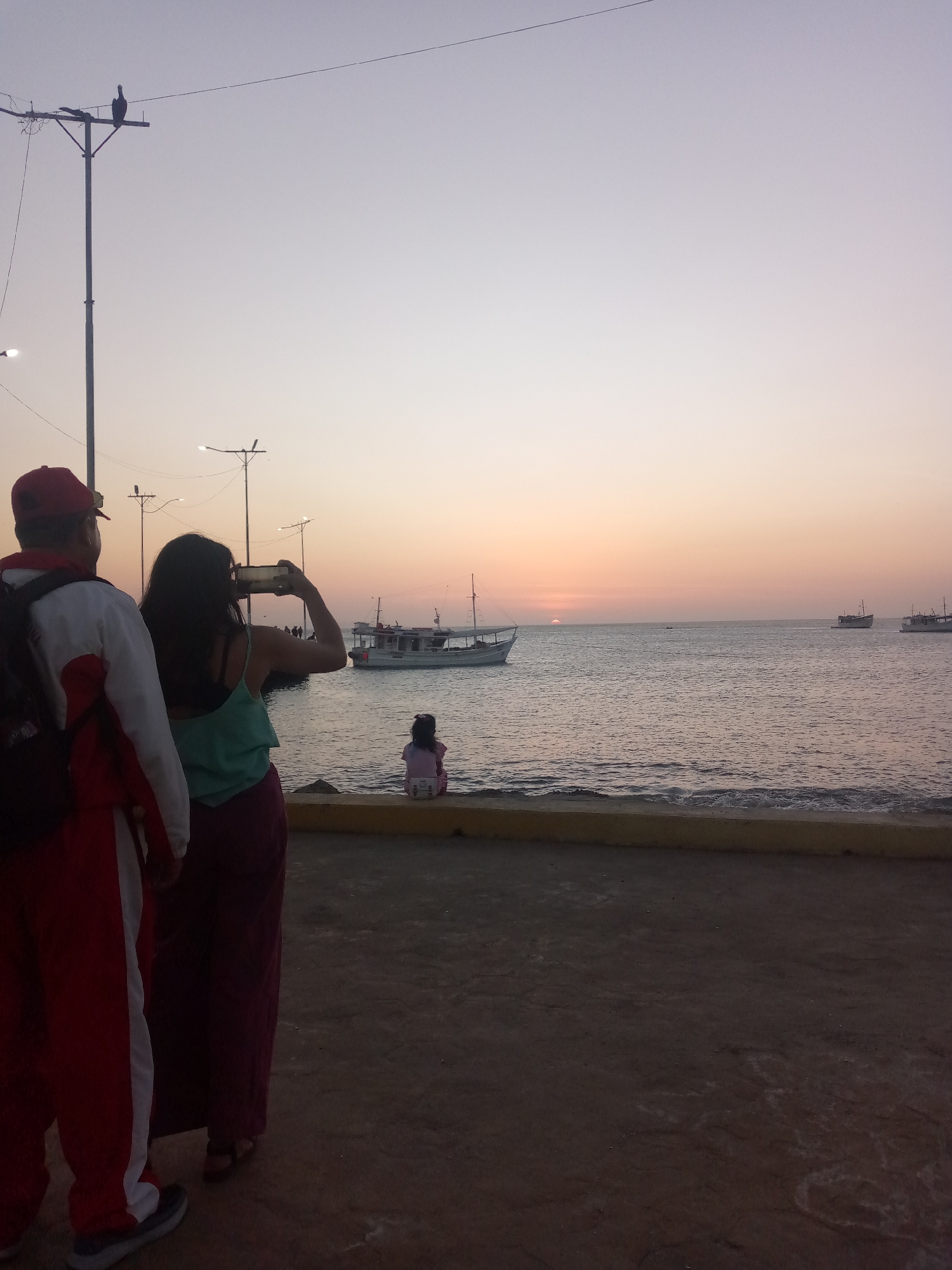 The image depicts a sunset over the sea in Juan Griego, Venezuela. In the foreground, a man wearing a red and white tracksuit stands next to a woman who is holding a smartphone up to take a picture of the sunset and a boat in the distance. In the midground, a young girl is sitting on a low wall by the sea. Additional boats are visible on the horizon, and streetlights are scattered throughout the scene. The sky is a mix of orange, yellow, and light gray hues.