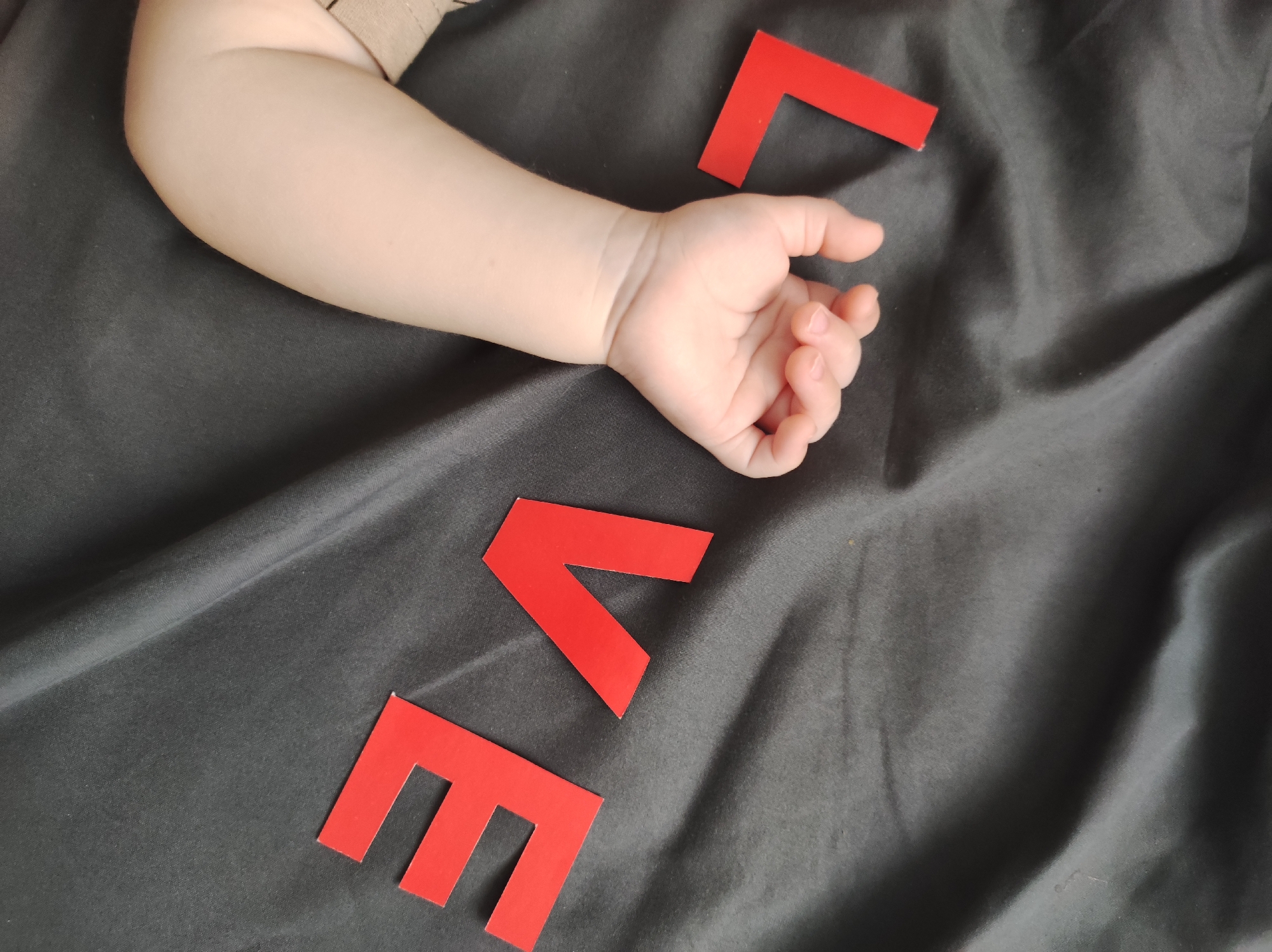 The image shows a baby's arm and hand resting on a dark gray, wrinkled fabric. Red, block-letter cutouts, spelling out "LOVE", are arranged near the baby's hand on the fabric.
