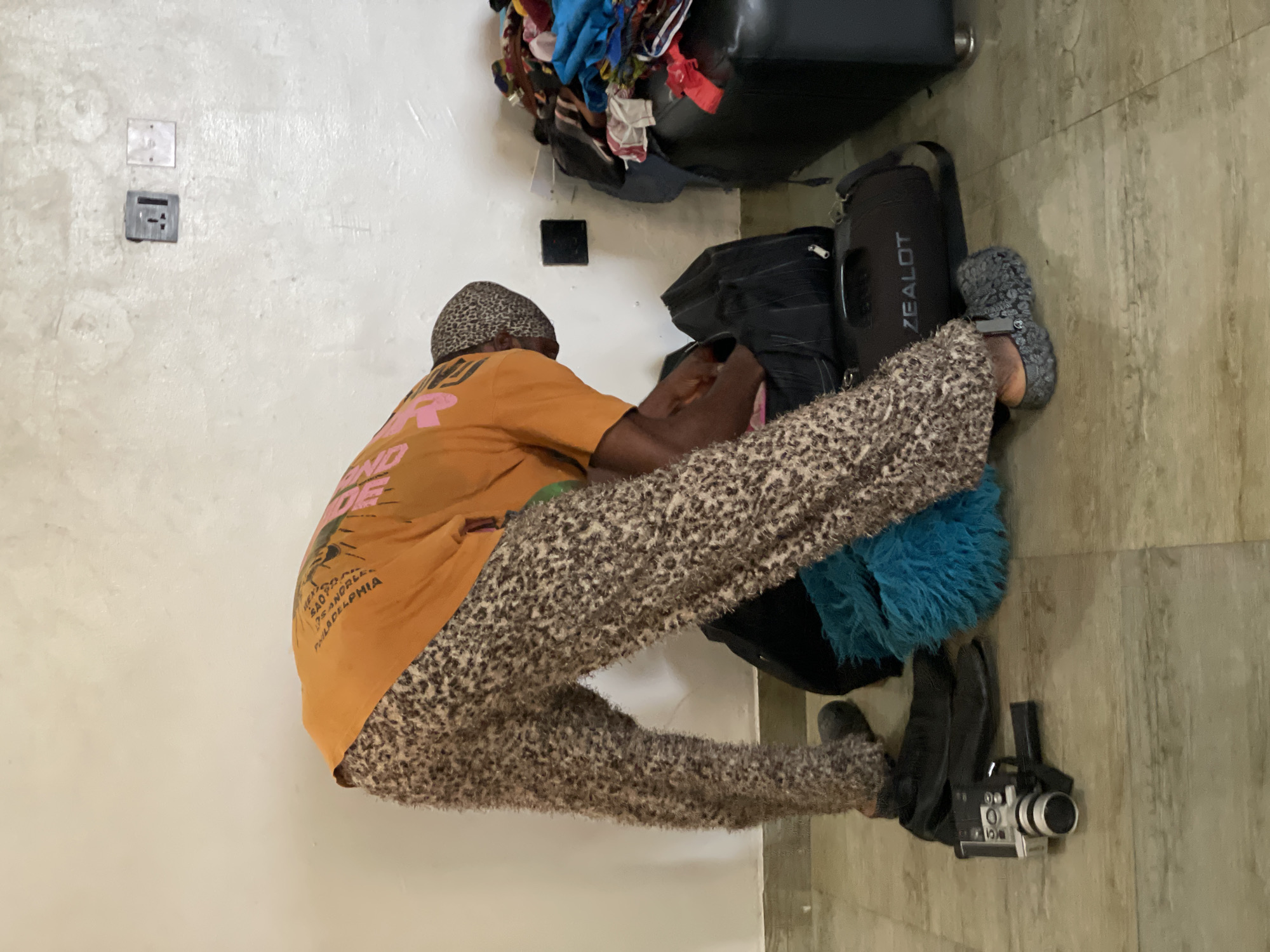 A person wearing a leopard print bandana, an orange t-shirt with pink lettering, and fuzzy leopard print pants is bent over packing items into a black duffel bag. The person is wearing gray Crocs on their feet. To the left of the person, there are a pair of black shoes and a silver video camera. Behind the person, a large black bag with the word "ZEALOT" is visible, as is a bright blue fuzzy object. To the right, a black leather couch is piled with various colorful clothes. The scene appears to be indoors, with a white wall in the background that has two electrical outlets. The floor is tiled in a light brown wood-grain pattern. This scene is likely located in Ado, Nigeria, based on context not visible in the image.