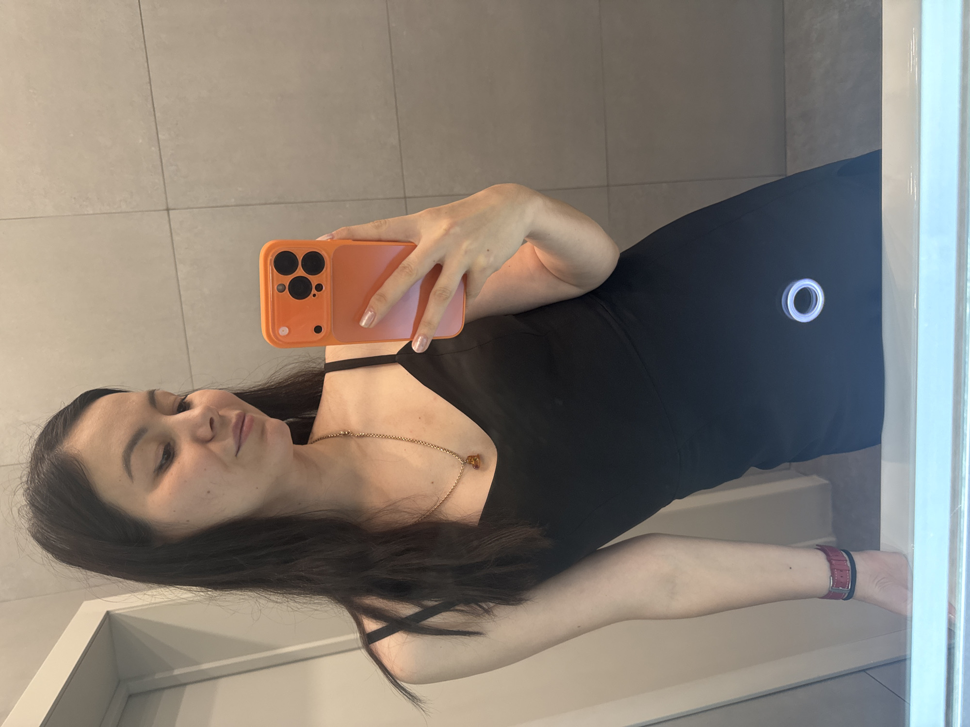 A young woman with long, dark hair and light skin is taking a selfie in a bathroom. She is wearing a black slip dress and a gold necklace with a heart-shaped pendant. Her right hand is holding an orange iPhone with a pink gradient case. The bathroom has beige tiled walls and a white countertop. The woman is looking down at her phone, and her expression is soft. The image is taken from a slightly high angle.
