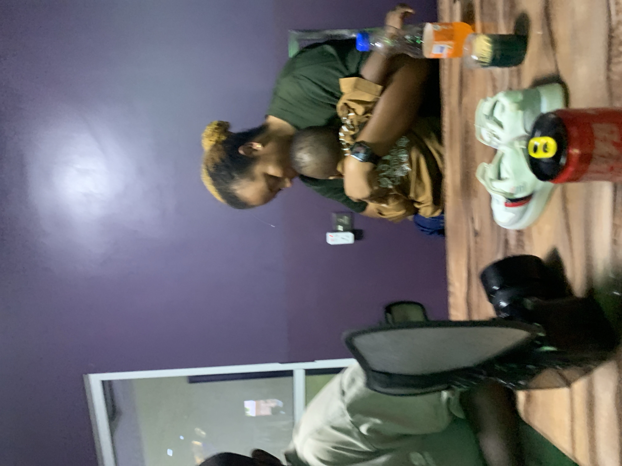 An indoor scene depicts two individuals, an adult woman and a child, in an embrace at a wooden table, with various objects in the foreground. **Individuals:** * An adult woman is seated on the right, facing left, with her head tilted downwards towards the child. She has light brown hair styled in a bun and wears a dark green short-sleeved top. A dark-faced wristwatch is visible on her left wrist. She is holding the child with her left arm. * A child, appearing to be a toddler or very young, is held by the woman, facing away from the camera, with its head resting against the woman's left shoulder. The child wears a brown long-sleeved shirt with light-colored patterns or text on the front. Its arms are wrapped around the woman's neck. * A third individual is partially visible on the far left, seated at the same table, with only a portion of a light olive-green long-sleeved shirt and an arm discernible, facing away from the camera. **Objects:** * On the wooden table in the foreground, from left to right: A black and grey studio light modifier (e.g., a small softbox or reflector) stands vertically. Next to it, a black camera body with a lens attached lies horizontally. Two pairs of light-colored (white/light grey) athletic sneakers are present, one pair featuring red and dark stripes on its side. A red beverage can with a yellow pull tab is visible. A clear plastic bottle contains an orange carbonated beverage. A dark green glass holds a yellow/green cylindrical object, possibly a candle or small container. **Environment:** * The background features a solid, matte purple wall. On the upper left, a white-framed glass partition or window reflects ambient light. A white two-socket electrical outlet is mounted on the purple wall to the left of the woman. The foreground consists of a wooden table with visible plank construction and prominent grain. The scene is illuminated by artificial light, indicated by a localized bright reflection on the upper-center portion of the purple wall. **Actions and Interactions:** * The woman is embracing the child, holding it closely, and looking down. The child reciprocates the embrace, holding onto the woman's neck. The individuals are situated at a table, suggesting a social or casual gathering. **Location Note:** * There are no specific visual cues within the image that confirm the location as the city of Jos, Nigeria. The
