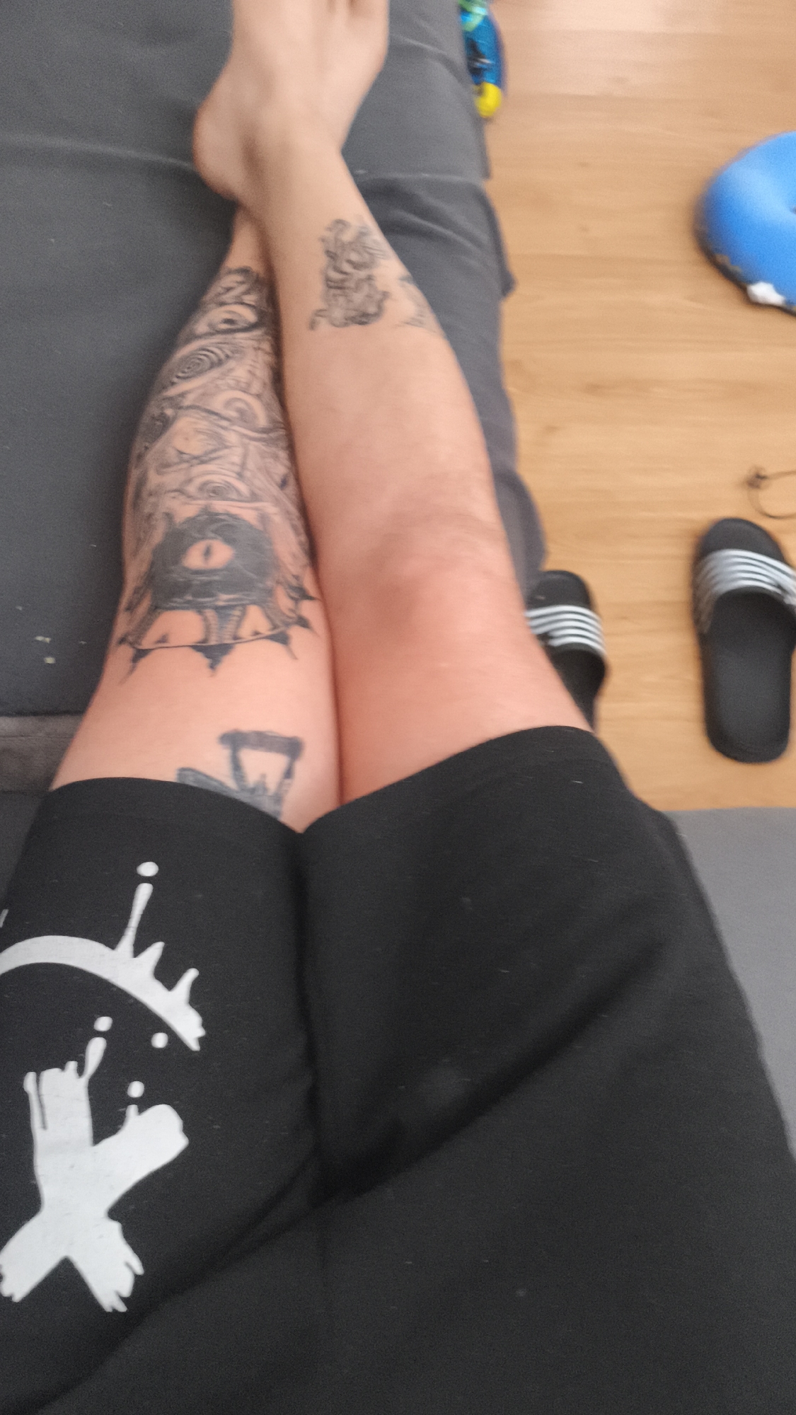The image shows a person's legs crossed while sitting on a gray couch. The person is wearing black shorts with a white graphic on one leg. One leg is heavily tattooed with a detailed black and white design, including an eye motif. The other leg has two smaller tattoos. The person's feet are visible, with one foot crossed over the other leg. Beside the couch, there is a wooden floor with a pair of black and silver Adidas-style sandals. A blue helmet is also visible. There is no information or context to determine that this scene is taking place in Nowy Tomyśl, Poland.