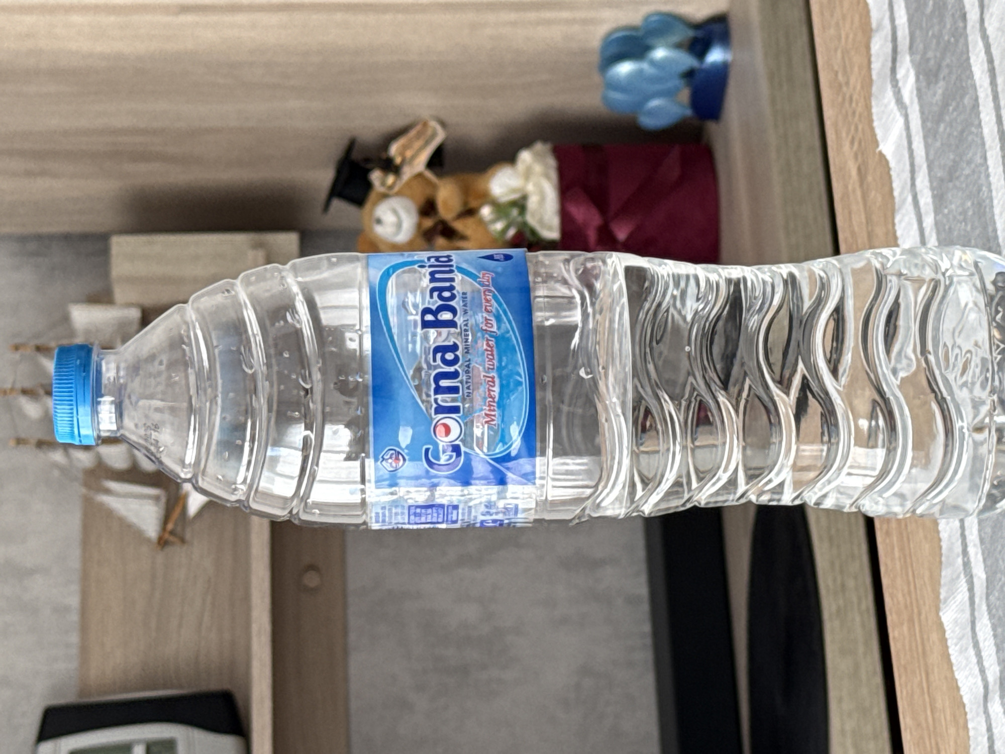A clear plastic bottle of "Gorna Bania" natural mineral water is lying horizontally on a surface. The label is predominantly blue and white. The cap of the bottle is blue. The background includes various items: a small boat model, a graduation bear, a red decorative item, a blue item, a piece of furniture, and a striped fabric. The location is Lovech, Bulgaria, as "Gorna Bania" is a brand of mineral water from Bulgaria.