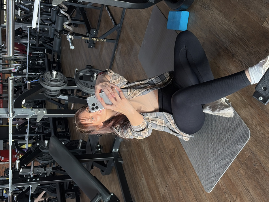 A female subject is observed seated on a grey yoga mat on a wooden floor within a gym. She holds a light grey smartphone with both hands, extended forward, capturing a selfie. The subject wears a plaid shirt unbuttoned over a light-colored crop top, black athletic leggings, and white athletic shoes with white socks. Her hair is pulled back with bangs. To the right of the yoga mat is a blue yoga block. The background reveals numerous black gym machines, including barbell racks with weight plates, benches, and other strength training equipment. Russian text, "СПОРТ ПЕРЕПОНОИ," is visible on a piece of gym equipment, indicating the location as the Sport Pereponov Fitness Club in Chaikovsky, Russia. Blurred figures of other individuals are discernible in the deep background.