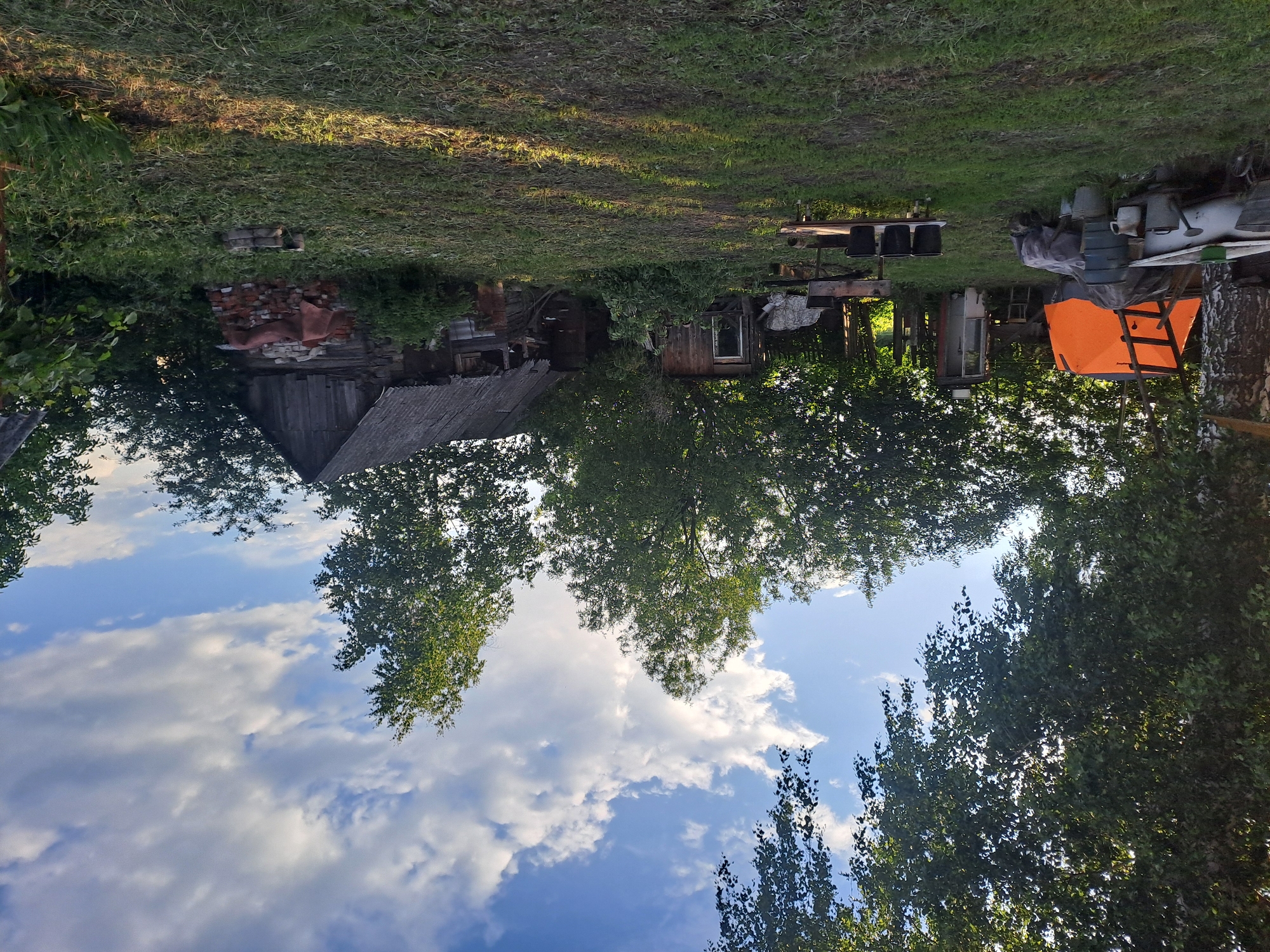 This image captures a rustic, outdoor scene, likely a private plot or backyard in a rural area like Полянки, Russia. The sky above is partially cloudy with large white cumulus clouds set against a blue backdrop, suggesting fair weather. Lush green trees, with their leaves illuminated by sunlight, frame the upper and lower parts of the scene (when correctly oriented).
The ground is covered in grass, showing varied textures and tones, with areas brightly lit by what appears to be late afternoon or early evening sun, casting long shadows. Several weathered wooden structures are present: a larger building with a sloped, possibly corrugated roof on the left, and a smaller shed with a visible window further to the right. Piles of red bricks and other discarded materials are stacked near the larger building, indicating ongoing work or storage.
Prominently featured on the right side of the image is an orange tent-like structure or tarp-covered shelter, with some wooden planks or a ladder leaning against it. Various utilitarian items, such as metal buckets, pots, and a makeshift table with dark containers, are scattered around the grounds, suggesting active use or storage in this lived-in, somewhat cluttered environment. No explicit activity is taking place, but the scene conveys a sense of a working, rural homestead. There is no visible text in the image.