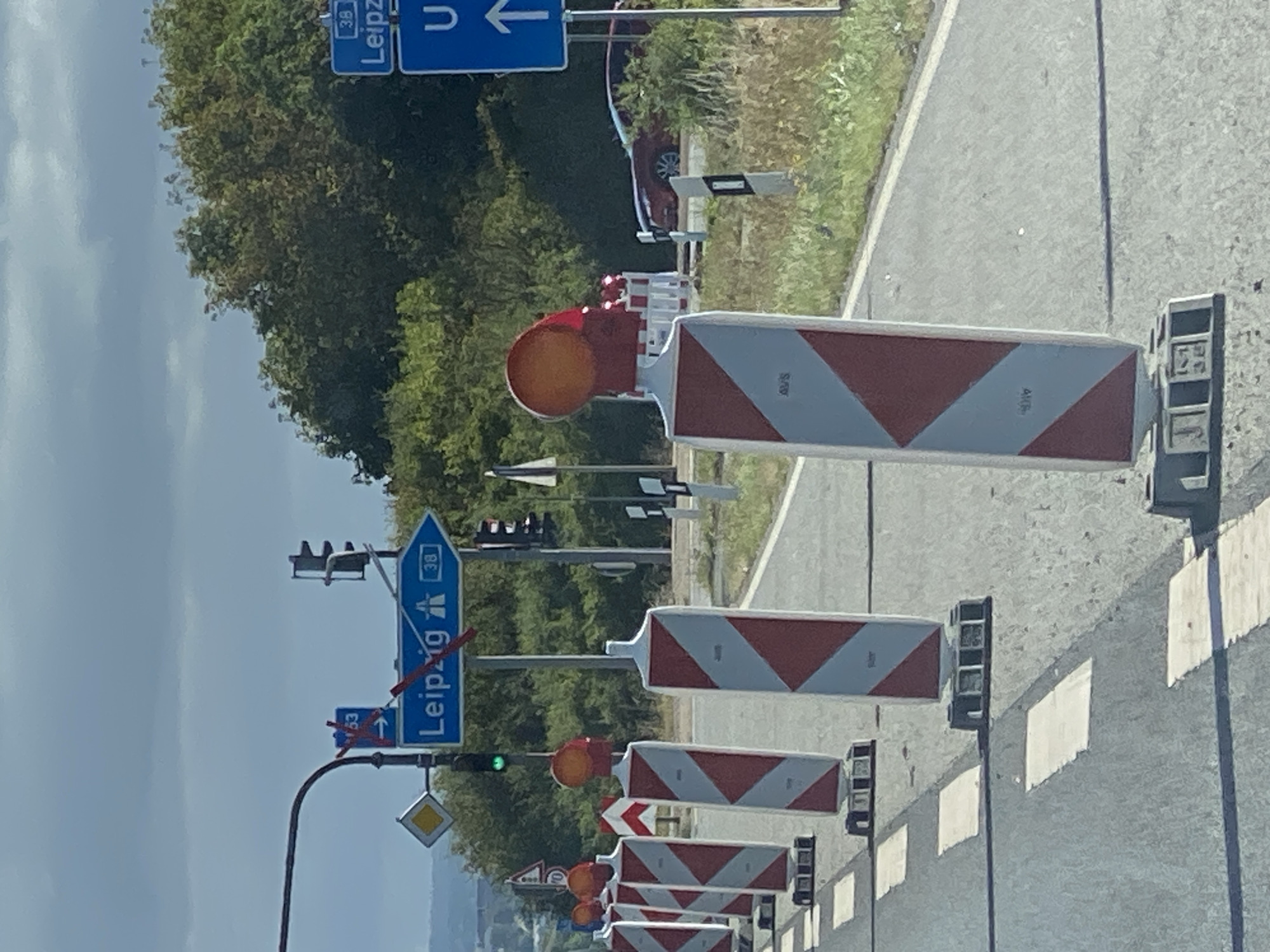 The image depicts a roadside scene with construction barriers. In the foreground, several orange-topped traffic pylons with red and white diagonal stripes are lined up on the asphalt road. Beyond the pylons, a blue directional sign points to the right, indicating "Leipzig" with a route number "38" and an icon of an expressway. Above this sign, a smaller blue sign shows "63" with an arrow pointing left. A traffic light is visible, showing a green light. In the background, trees and foliage form a dense screen, and a red car is partially visible on the right side of the road. The sky is overcast with clouds. The location is identified as Werther, Germany, with the specific context of road construction or lane closure.