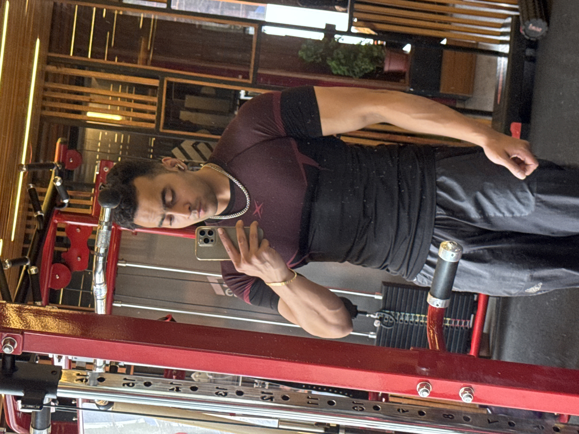 A young South Asian man is taking a mirror selfie in a gym. He is wearing a dark maroon and black t-shirt, dark gray sweatpants, and a silver chain necklace. He has short dark hair styled in a bun. The gym appears to have wooden accents on the walls and floor, with gym equipment visible, including a weight machine and a foam roller. The scene suggests a workout session is in progress or has recently concluded. There is no indication of the specific city or country.