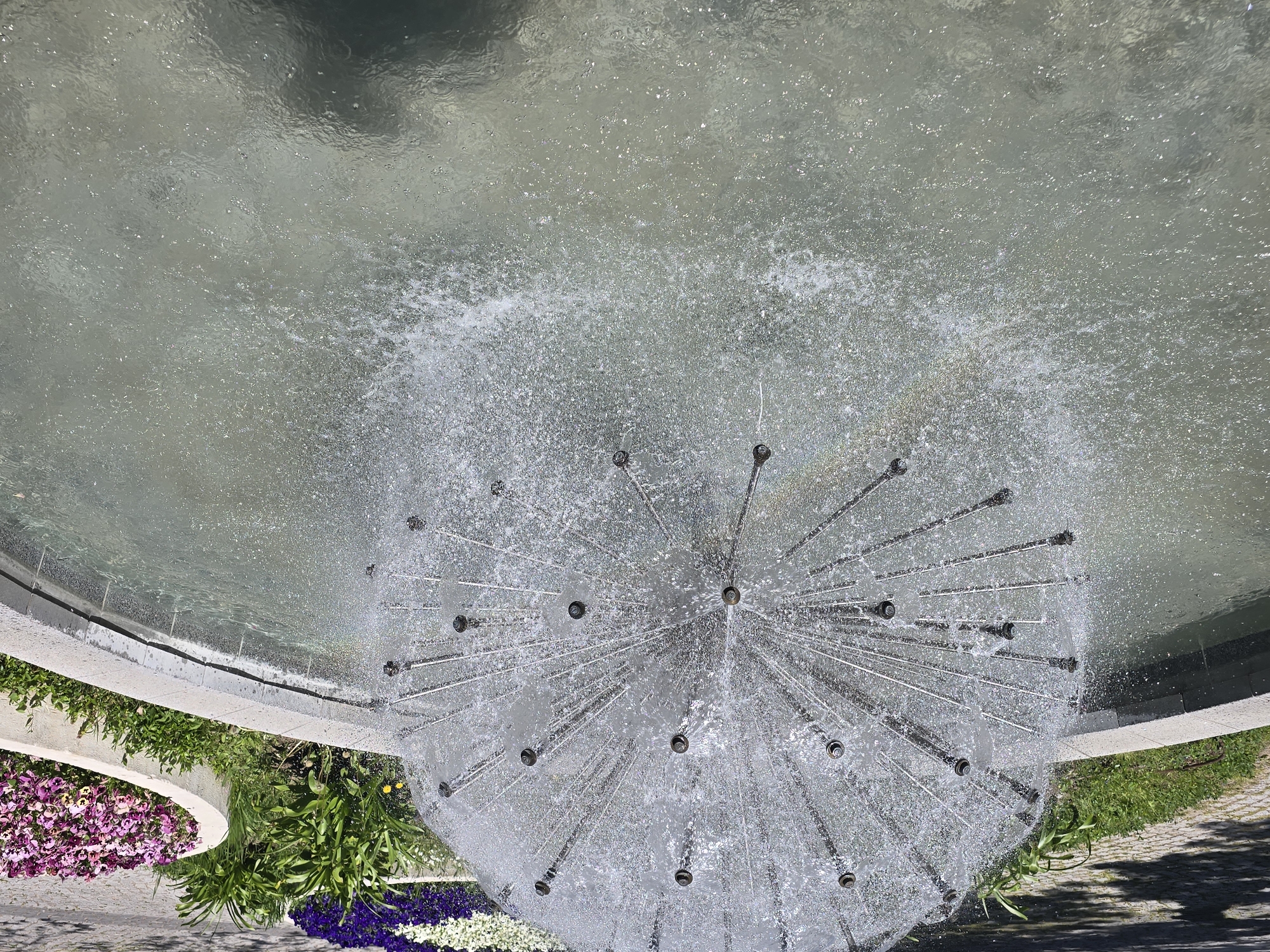 The image is a high-angle shot of a fountain in Unknown, Unknown. The fountain is circular, filled with water, and has a central structure composed of multiple metal rods radiating outwards with nozzles that spray water. The surrounding area features various types of flowers and greenery. A concrete edge surrounds the fountain. The image is well-lit, indicating daytime conditions.