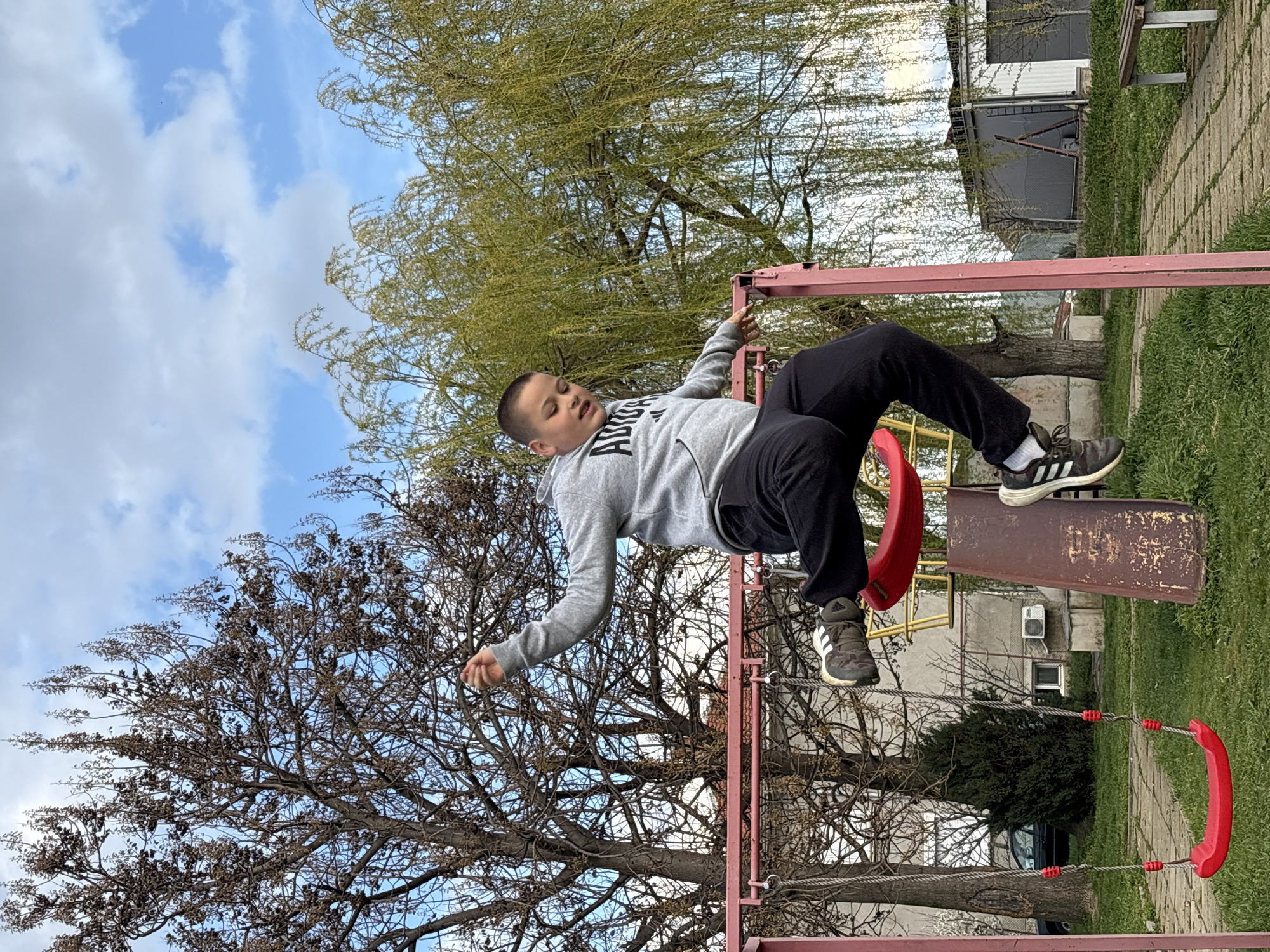 A young boy is upside down on a playground swing in an unknown city. He is wearing a gray hoodie and black pants. His Adidas sneakers are visible. He is holding onto the swing set frame. The environment includes trees with budding leaves, a blue and cloudy sky, a grassy area, and a concrete pathway. In the background is a building. The scene appears to be outdoors in a public park.