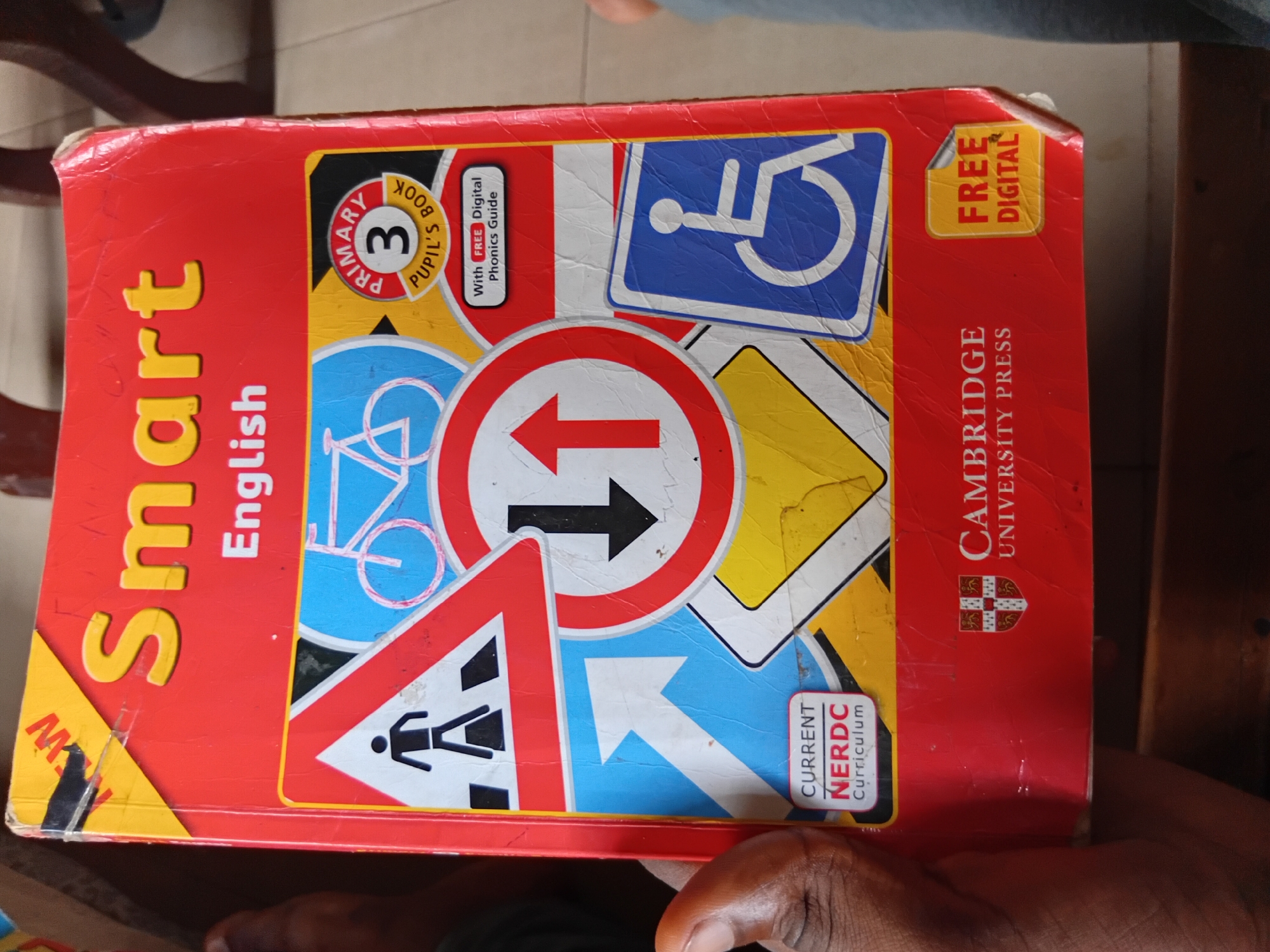 The image shows a "Smart English Primary 3 Pupil's Book" with a red cover adorned with various road signs, including a pedestrian crossing, a bicycle, a one-way street sign, and a wheelchair-accessible symbol. The book is being held by a person's left hand. In the bottom right corner, there is a yellow sticker that says "FREE DIGITAL." Below the book cover, the logo of "Cambridge University Press" is visible. The scene is set in Nkpor, Nigeria.