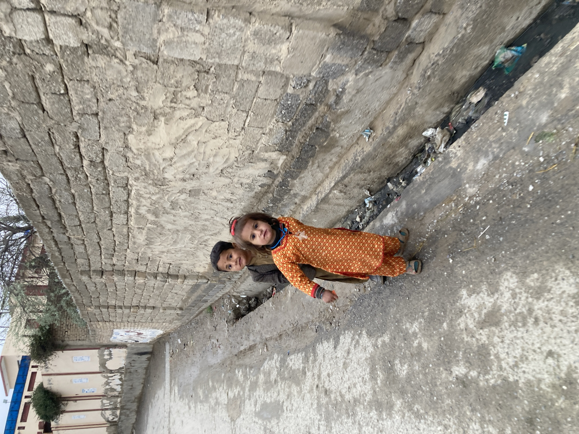 Two children stand on a concrete street next to a brick wall, facing the camera. The girl wears an orange patterned outfit with a red headband and blue necklace. The boy wears a brown jacket and stands slightly behind her. A narrow, trash-filled drainage ditch runs alongside the street. The scene is in Quetta City Tehsil, Pakistan, judging from the setting and appearance of the children.