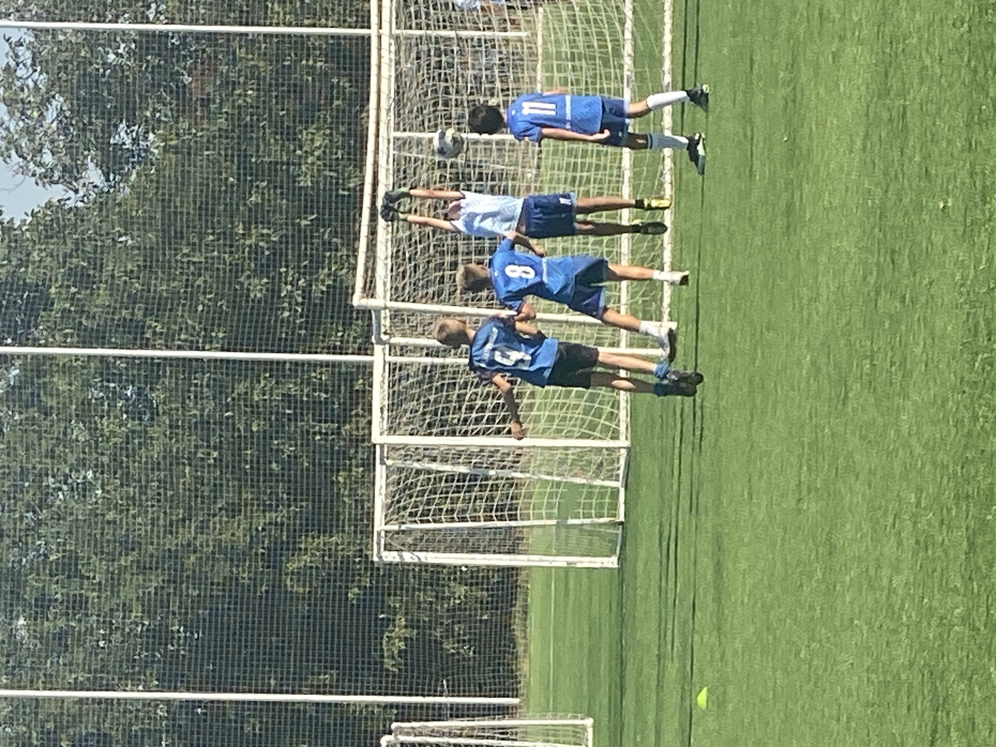 The image shows a group of young boys playing soccer on a green field with soccer goals and nets in the background. The boys are wearing blue soccer jerseys and shorts. One boy in the center is jumping up, reaching for a soccer ball. Another boy is looking up at the ball, and two other boys are watching. The scene is outdoors on a sunny day. The provided metadata indicates this image was taken in Krasnodar, Russia.