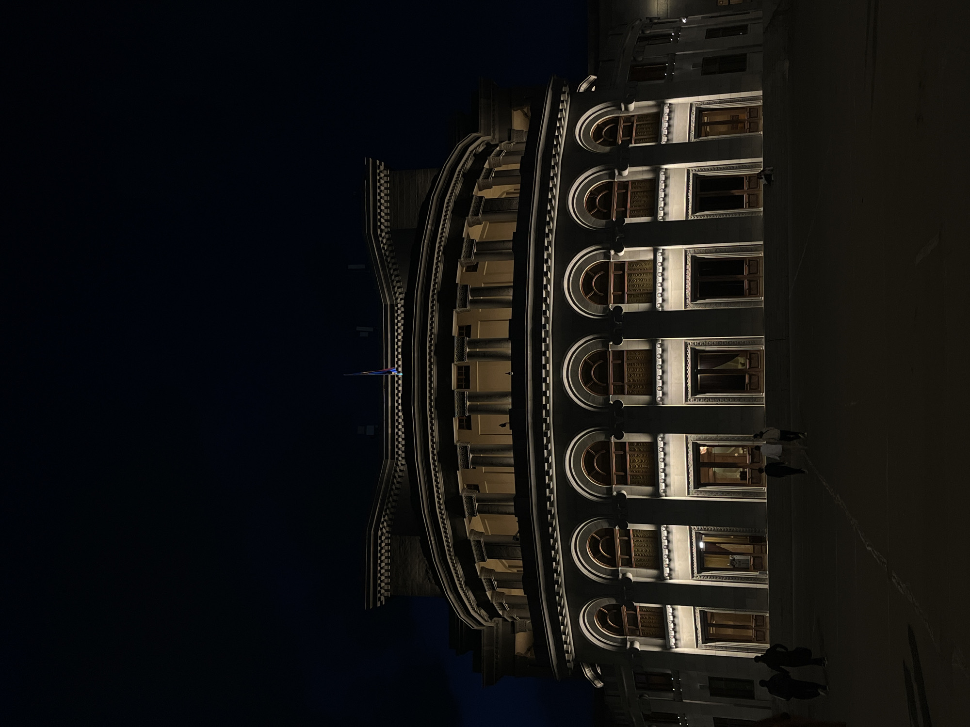 The image shows a building facade at night in Yerevan, Armenia. The building is brightly illuminated and features a curved architectural design with numerous windows. Some people are walking in front of the building. The sky is dark.