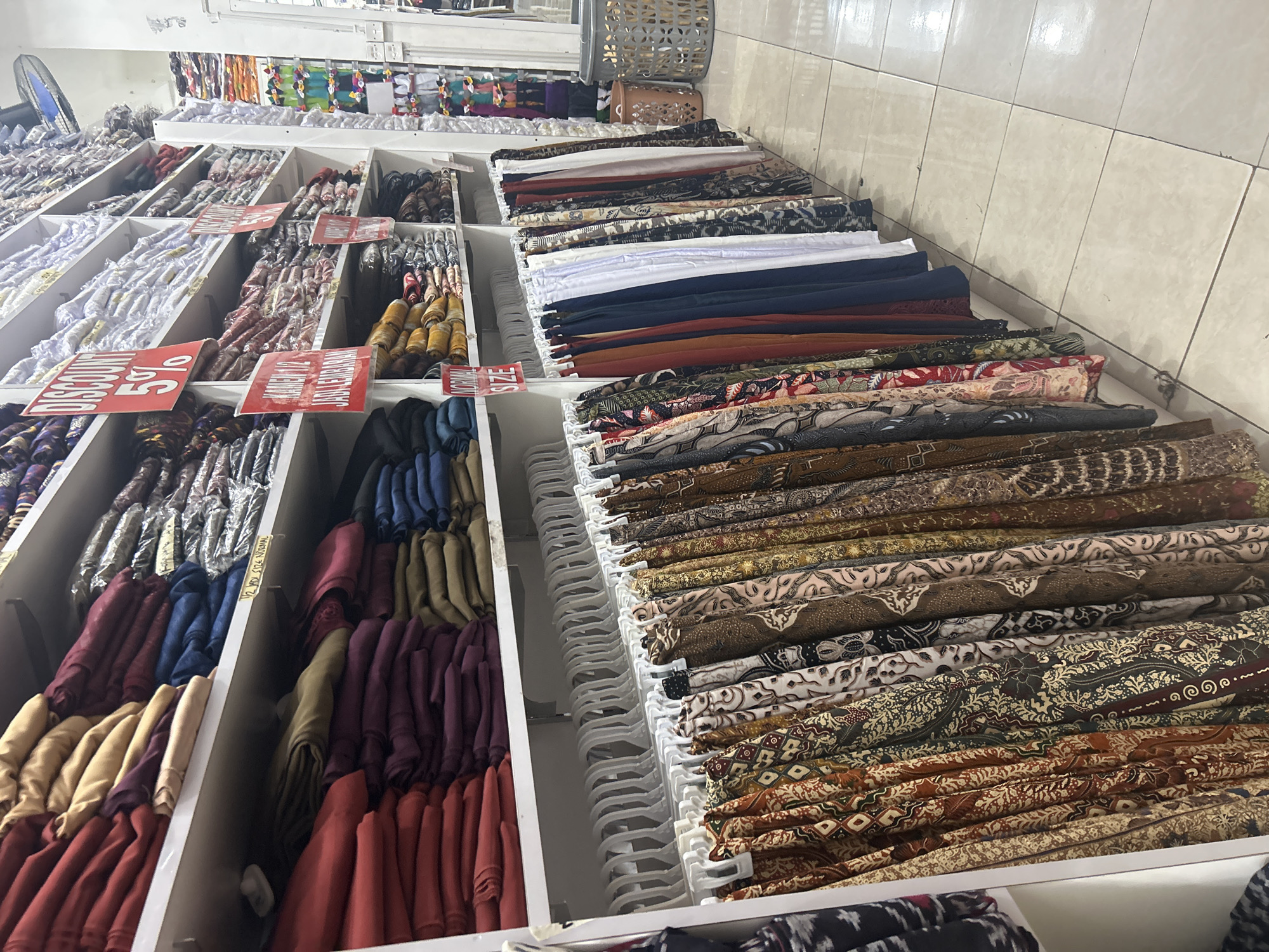 The image depicts a vibrant market stall in Ubud, Indonesia, filled with a wide variety of fabrics. The scene is set indoors, likely a shop or market, with white tiled walls and shelves overflowing with textiles. The primary subjects are the fabrics themselves, displayed in neat stacks and rolls. They exhibit diverse patterns, including traditional batik designs with intricate motifs, floral prints, and solid colors in shades of red, maroon, yellow, blue, and white. Many of the fabrics are folded and presented on white hangers, creating a visually appealing retail display.
Red discount signs are strategically placed among the fabrics, indicating sales and promotions. One sign clearly reads "DISCOUNT 50%". Another sign, partially visible, seems to indicate "NORMAL SIZE". The lighting suggests it could be daytime, typical for a market setting, with the overall atmosphere conveying a sense of abundance and commercial activity. The detailed patterns on the fabrics, characteristic of Indonesian textiles, are a notable detail, hinting at the cultural richness of the location. The lack of people in the frame focuses attention solely on the merchandise, highlighting the array of colorful and patterned fabrics available for purchase.