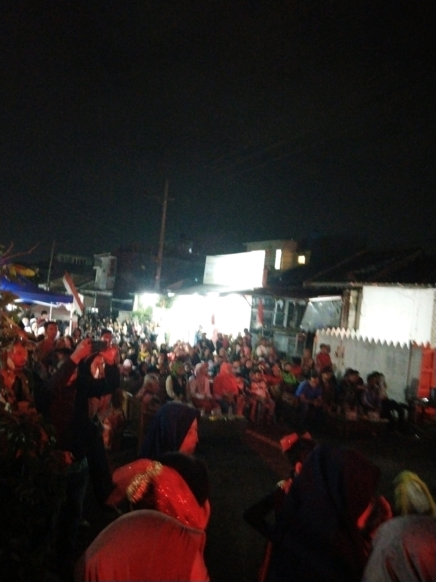 The image depicts an outdoor gathering at night in Singosari, Indonesia. A large crowd of people, many wearing headscarves, are visible, suggesting a community event or celebration. The scene is illuminated by artificial lights, casting shadows and highlighting the faces of some individuals. In the background, buildings and utility poles are discernible. A red and white flag, possibly the Indonesian flag, is also present, adding a patriotic element to the scene. The overall atmosphere appears festive and communal.