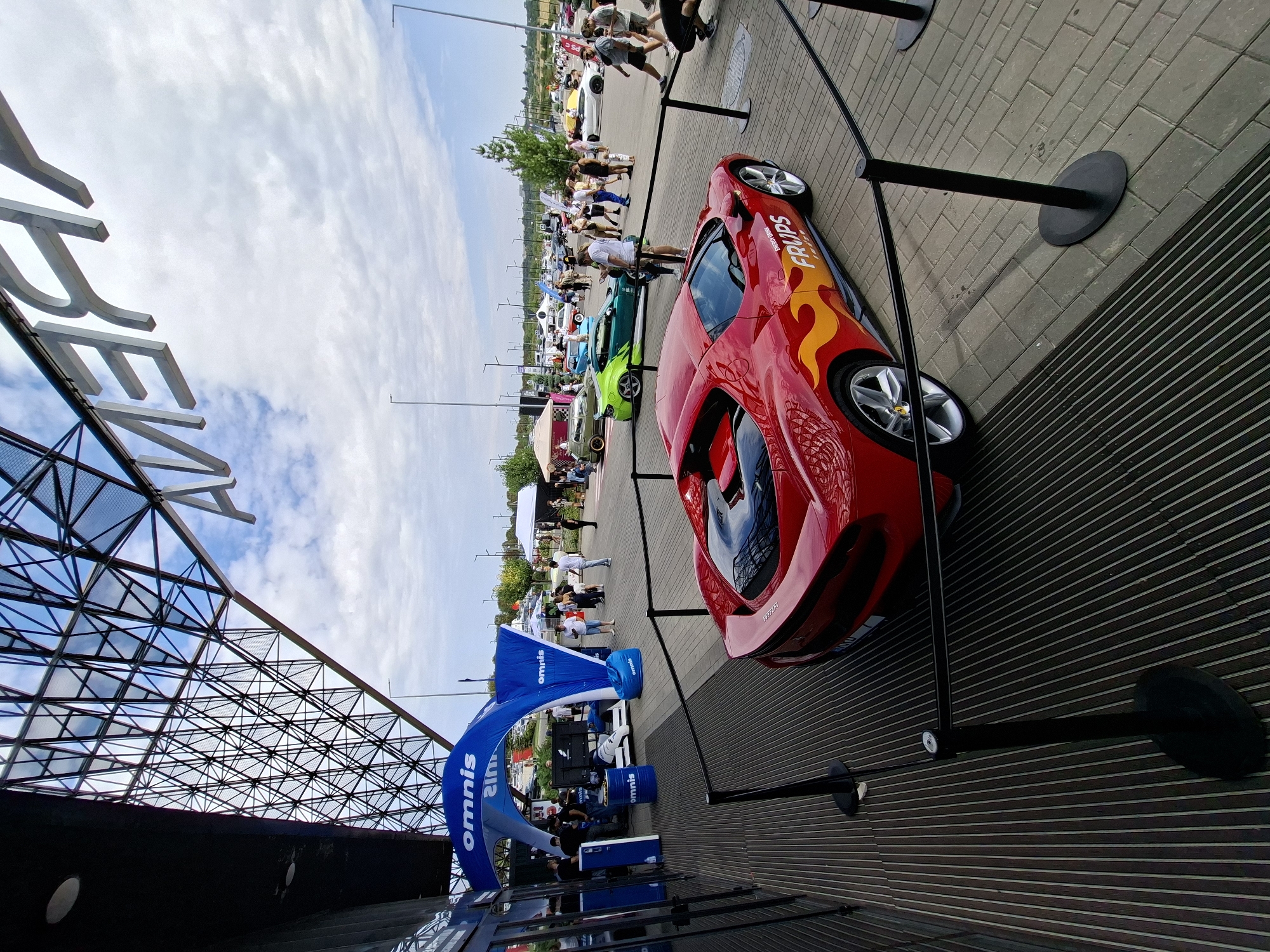 The image depicts an outdoor car show in Stăuceni, Moldova, likely held at an exhibition center given the prominent "ARENA" signage visible on the building's facade. In the foreground, a vibrant red sports car with flame decals and "FRUPS" branding is displayed behind a black velvet rope barrier. The car is positioned on a paved area. To the left, a blue inflatable archway with "omnis" branding stands over a display area featuring booths and people interacting. Several other cars, including a bright green modified vehicle, are visible further in the background. A crowd of people are milling around the various exhibits and vehicles. The sky is partly cloudy, and the scene suggests a daytime event.