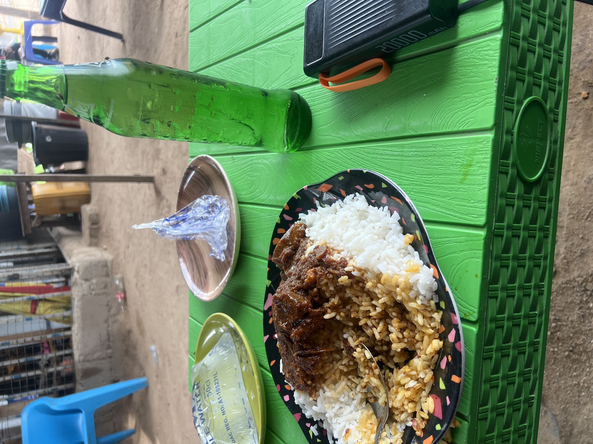 The image shows a meal on a green plastic table. The meal consists of rice and meat, served on a black plate. Beside the plate is a green soda bottle, a power bank, and two small plates covered in cellophane or plastic. The environment appears to be outdoors, potentially in Jalingo, Nigeria, given the context, though there are no specific landmarks or signs to confirm the location. A blue plastic chair is visible in the background. The table has a woven plastic texture.
