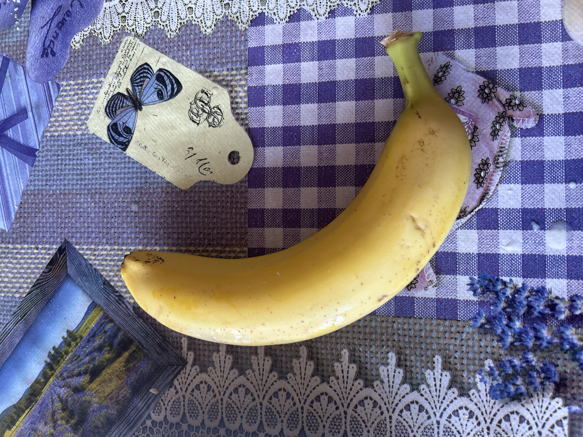 The image depicts a yellow banana lying on a surface covered with various purple and white patterned materials, including a gingham check cloth, lace, and a textured fabric. A framed picture of a lavender field and a tag with a butterfly illustration are also present. Based solely on the content of the image, there is no indication of the location being Dve Mogili, Bulgaria, or of any people, actions, or interactions.