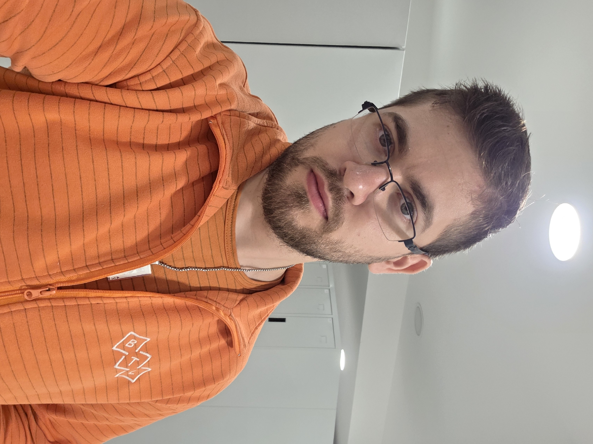 A person wearing glasses and an orange, horizontally striped zip-up hoodie with a logo near the zipper is shown in an indoor setting. The person is positioned at a slightly angled perspective, with the camera pointing upwards. The background features white walls and what appears to be office cabinets. There is a ceiling light visible in the upper right corner. It is impossible to determine the location in the city of Plovdiv, Bulgaria.