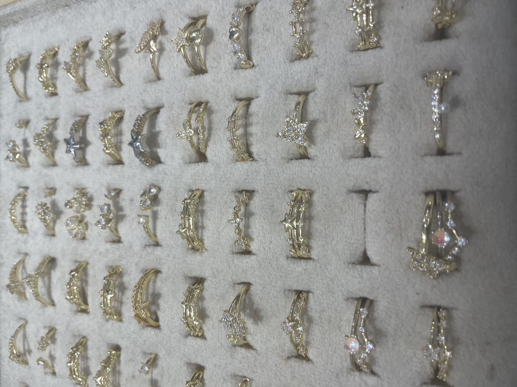 This image shows a top-down view of a display case filled with gold rings. The rings are arranged in rows and columns. Each ring has a unique design, with some featuring small diamonds or other gemstones, while others are plain gold bands. The rings are displayed on a textured, grey background that appears to be velvet or felt. The display case is well-lit, and the rings are in focus, showcasing their intricate details. The image does not contain any information about the city of Yunus Emre, Turkey, nor does it depict any people, actions, or interactions.