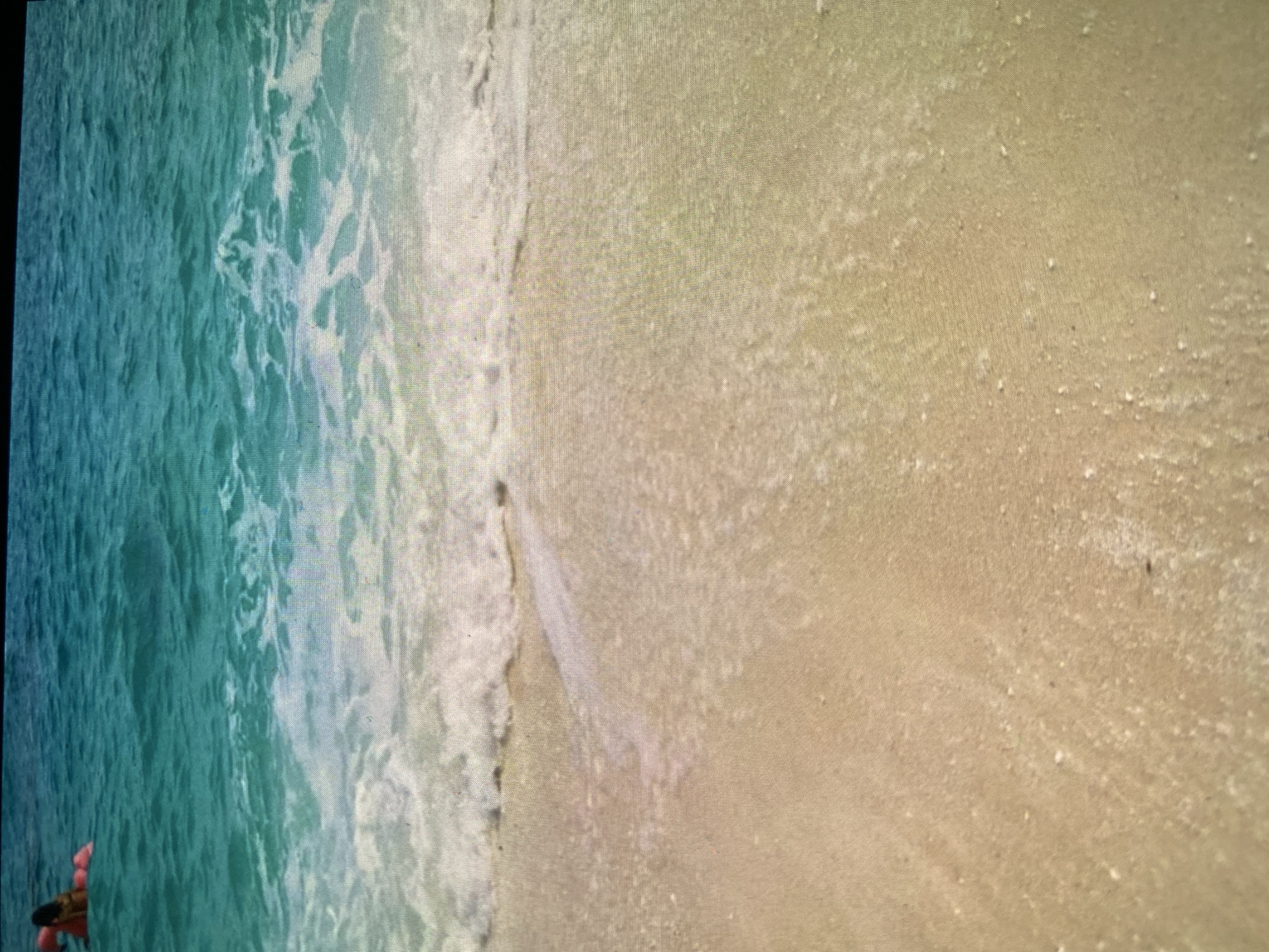 The image displays a vibrant beach scene captured from a high-angle or overhead perspective. On the left side, clear, turquoise-blue water, indicative of an ocean or sea, features white foamy waves breaking and receding towards the shore. The water’s color deepens further from the edge and transitions to a lighter, frothy white where it meets the land.
The right side of the frame is dominated by light golden-beige sand, exhibiting a fine, granular texture. The sand appears damp close to the water and gradually becomes drier further away, with small, scattered specks or shell fragments visible on its surface. A distinct line marks the interaction between the water's edge and the sand.
In the bottom-left corner, a person's hand or arm, adorned with a reddish garment, is partially visible, holding a dark object that could be a smartphone or a small camera. This suggests the person might be capturing the scene.
The lighting is bright, indicating a daytime setting. There is no visible text in the image. The image itself appears to be a photograph of a digital screen, showing a noticeable pixel grid across the entire scene. The depicted location is clearly a beach, which contradicts the provided context of Bankya, Bulgaria, an inland town.