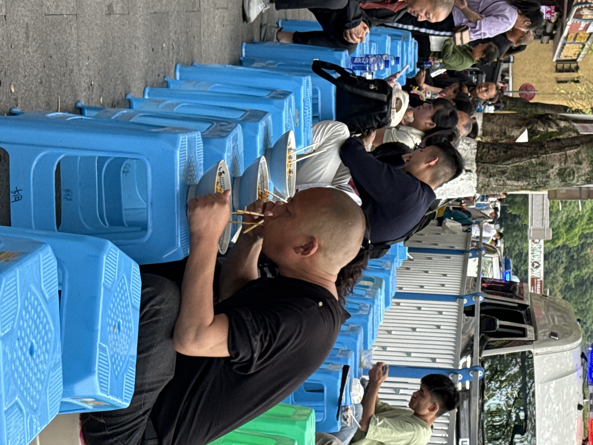 This vertical image depicts a bustling outdoor street food scene, likely in Yuzhong District, China. Numerous people, predominantly men, are seated on bright blue plastic stools, which also double as small tables for their meals. The scene is tightly packed, with rows of individuals lining what appears to be a sidewalk or an open-air eating area.
The primary activity is people eating. Many are shown consuming food, most likely noodles, from white and blue bowls using chopsticks. One man in the foreground, wearing a black T-shirt, is clearly visible holding chopsticks to his bowl. Others further down the line are also engaged in eating, conversing, or waiting.
Notable objects include the ubiquitous blue plastic stools, the bowls of food, chopsticks, and plastic water bottles, some of which bear the Chinese brand name "脉动" (Mài Dòng), a popular sports drink. One stool has a single Chinese character "怡" (yí, meaning 'happy' or 'pleasant') embossed on its side. In the background, there's a light-colored building, trees, and parts of a large vehicle, possibly a bus or truck, indicating an urban setting. The natural lighting suggests it is daytime, and the clear visibility implies fair weather. The atmosphere appears casual and lively, typical of a street food environment.