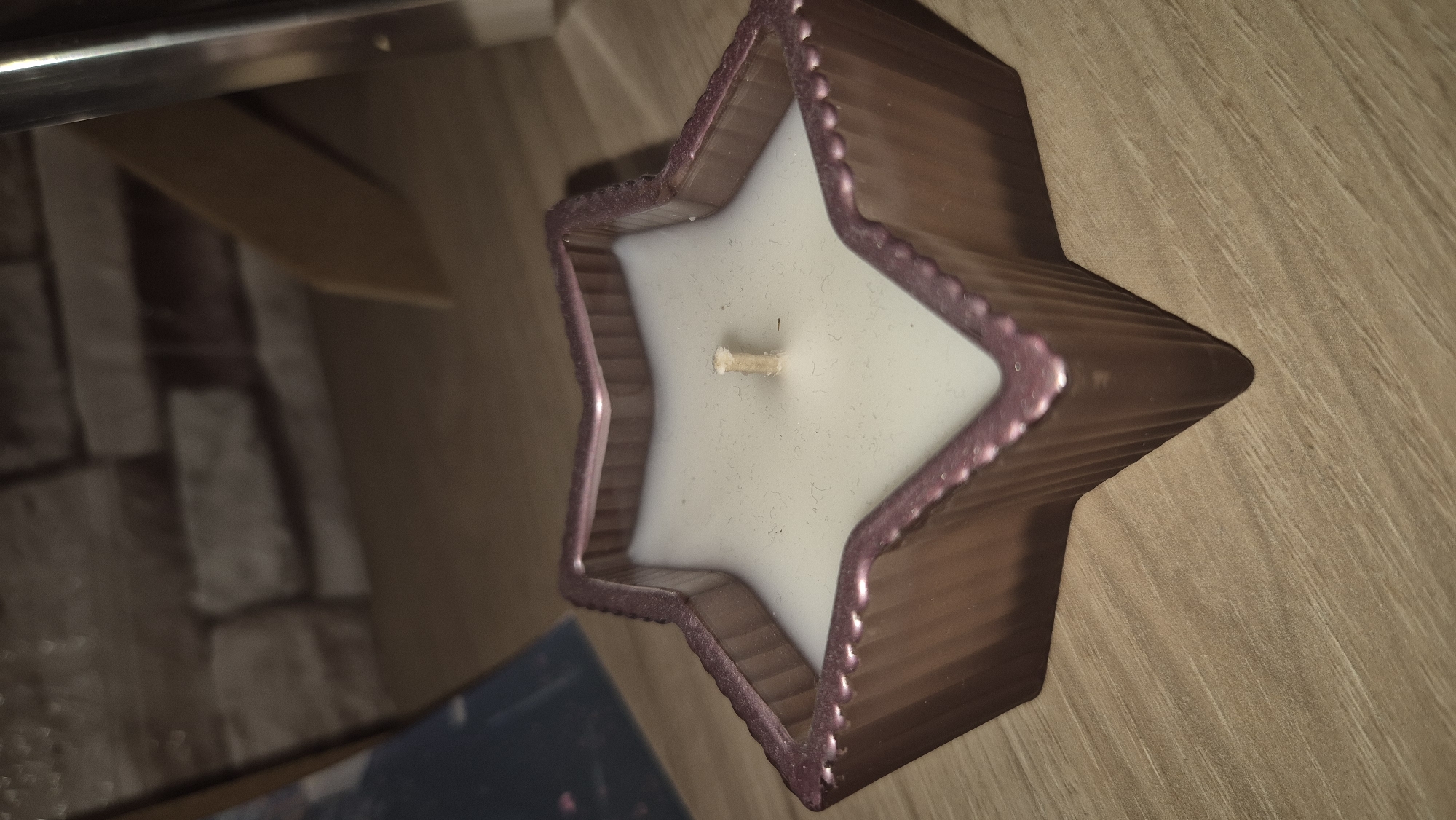 A star-shaped candle sits on a wooden surface. The candle is a pale white color and has a single wick in the center. The outer casing of the candle is a metallic rose gold color with ridged detailing. In the background, a brick wall is visible, along with a partially obscured picture frame. The lighting is dim.