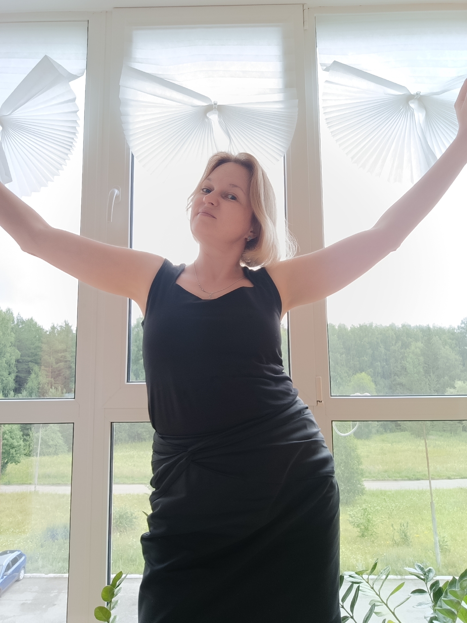 A woman with blonde hair is standing in front of a large window with white accordion-style blinds. She is wearing a black sleeveless dress and her arms are outstretched. Through the window, a green, tree-lined landscape is visible, suggesting a location outside of a city center. A blue car is parked in the distance. The overall lighting is bright, indicating it is daytime. The woman is looking at the camera with a slight smile and a raised eyebrow. The scene appears to be an interior of a room with a view of the outdoors, possibly an apartment or house in a suburban or rural area. The location within Марковский, Russia is not explicitly indicated by any visual cues within the image itself, but this is a context provided by the prompt.
