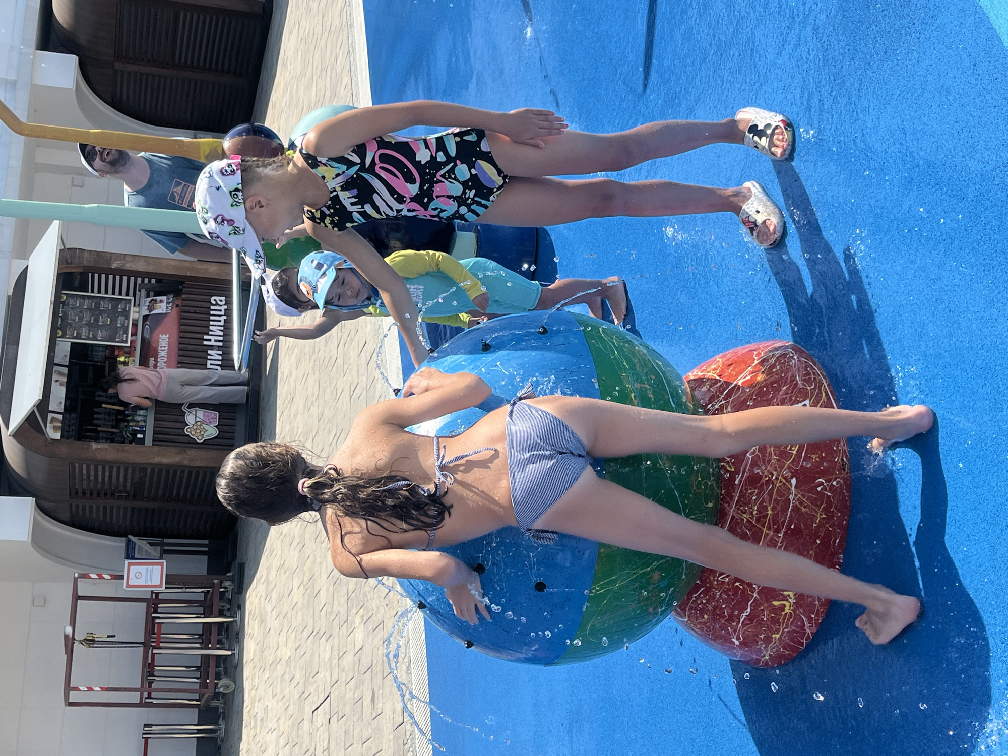 In Sochi, Russia, on a sunny day, two young girls are enjoying a water park. The scene is a bright blue splash pad with various water features. One girl, wearing a colorful swimsuit, is on her hands and knees, looking towards the camera. She has a playful expression. Beside her, a younger child in a yellow and blue rash guard is also on their hands and knees, facing away from the camera. They are both interacting with a large, inflatable globe that is spraying water. Another inflatable, red with white and yellow splatters, is partially visible to the right. In the background, a man in a dark t-shirt and sunglasses is visible, as well as a concession stand with striped awnings. The overall mood is one of fun and excitement on a warm day.