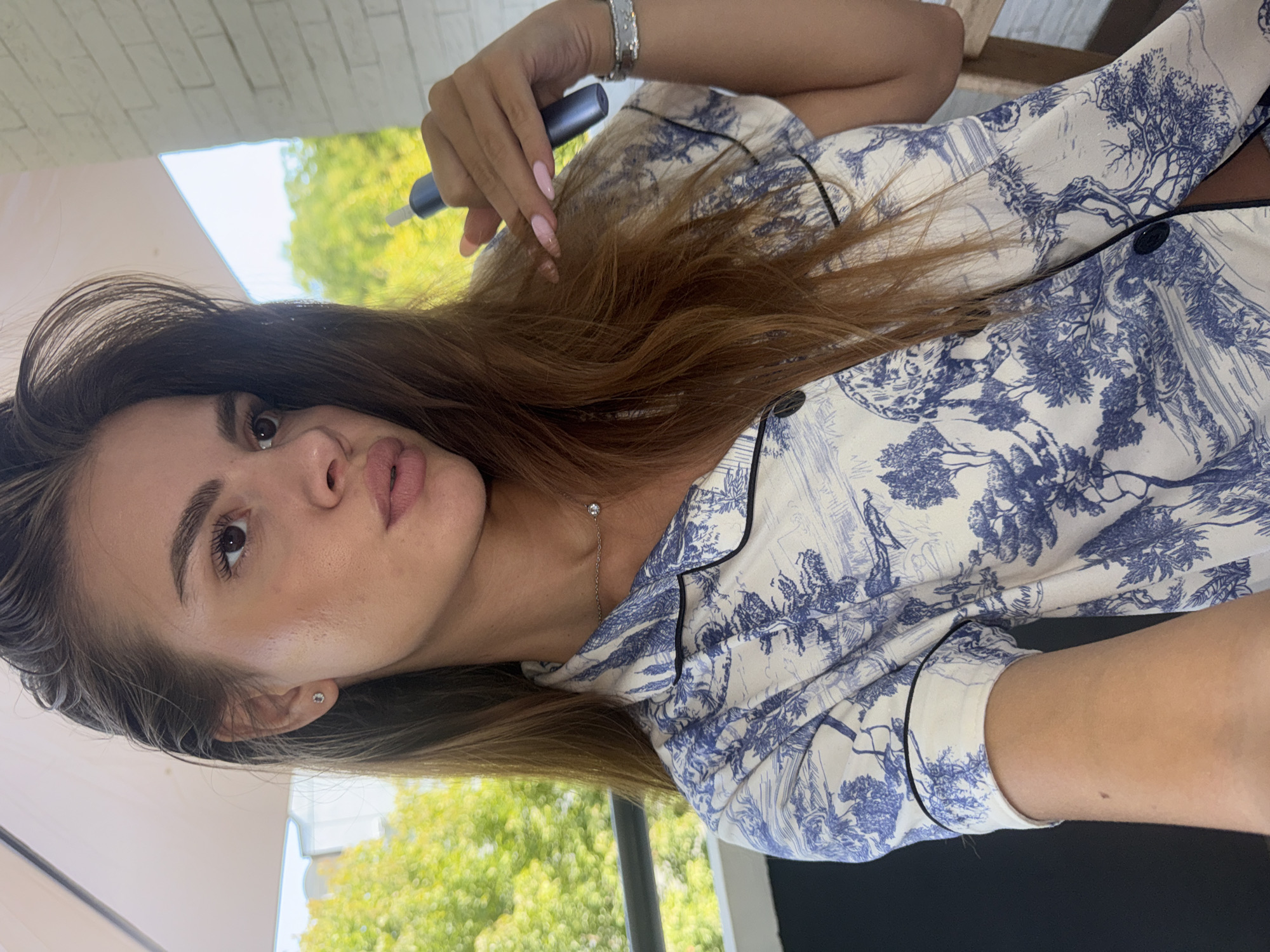 A young woman with long brown hair, wearing a white and blue patterned pajama top, is taking a selfie outdoors. She is looking to the right, and her right hand is holding a blue electronic device. She is also wearing a delicate silver necklace with a small pendant and a silver bracelet on her left wrist. The background shows greenery and a white brick wall, suggesting she is on a balcony or terrace. The image does not provide enough information to determine a specific location in the city of Plovdiv, Bulgaria.