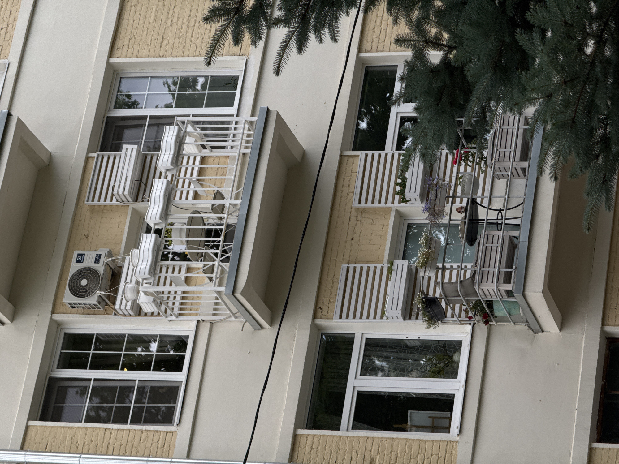 The image depicts the exterior of a multi-story building with yellow brick and beige facade, likely in Moscow, Russia. Two balconies are visible. The upper balcony has a white railing, an air conditioning unit mounted on the wall above it, and several white planters attached to the railing and wall, some containing plants. Two white chairs and a small table are also on this balcony. The lower balcony, positioned directly below the upper one, also features a white railing and a similar set of white planters on the wall and railing, holding plants, including red roses. This balcony has a round black table with two chairs, and a wooden bench-like seating arrangement with a planter box on top. A dark green coniferous tree partially obscures the right side of the lower balcony. The windows are white-framed with multiple panes. The overall scene suggests a residential building with decorative elements on its balconies.