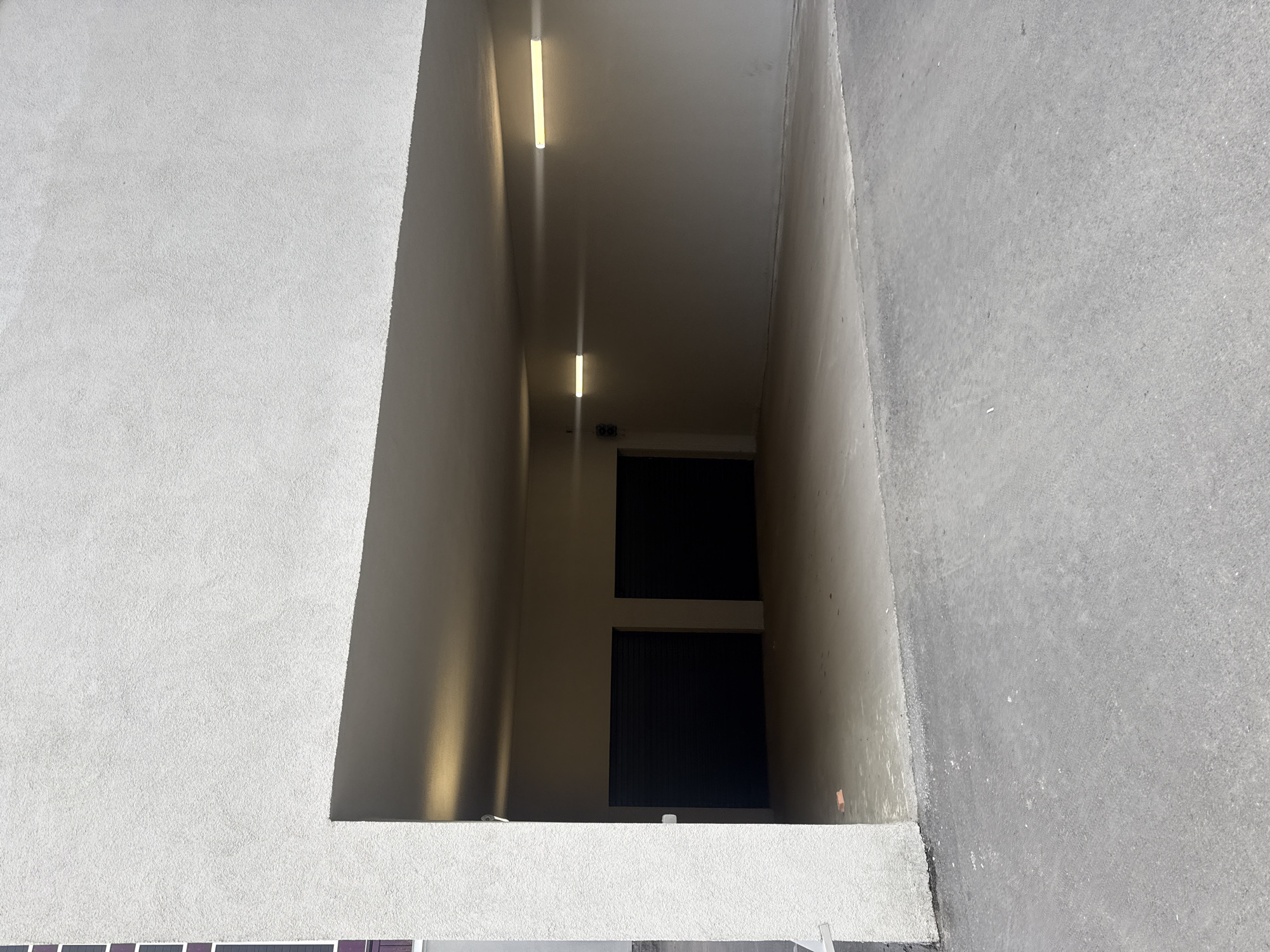 A low-angle, eye-level shot captures a narrow, recessed doorway or passage in a building. The walls of the passage are light gray and appear to be concrete or stucco. Two fluorescent light fixtures are visible on the ceiling of the passage. At the end of the passage, two dark doors or panels are stacked vertically, separated by a horizontal beam of light. The exterior walls surrounding the passage are textured, with one side appearing rougher than the other. The image lacks people, and the focus is primarily on the architectural elements. The location is unidentifiable from the image.