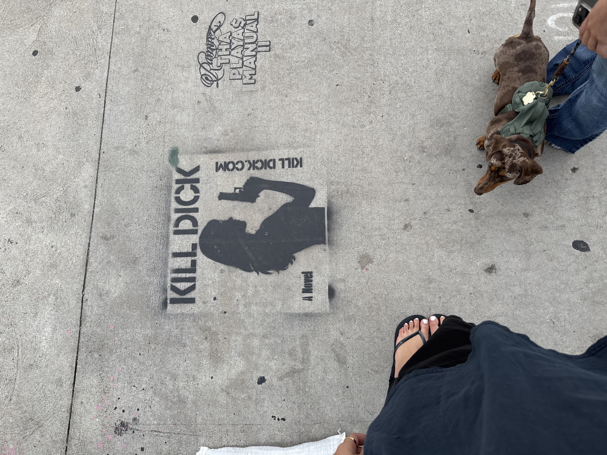 A top-down shot shows a sidewalk in Los Angeles, United States. On the concrete, stencil graffiti art depicts a woman holding a gun labeled "Kill Dick" and "A Novel," with the website "Kill Dick.com" written above. Nearby, another stencil reads "Tha Playas Manual." A brown dachshund wearing a green harness stands on the sidewalk, held on a leash by a person in blue jeans. Another person, wearing a long blue skirt and black flip-flops with white painted toe nails, stands near the "Kill Dick" stencil. The sidewalk shows typical wear and tear with a few black spots.