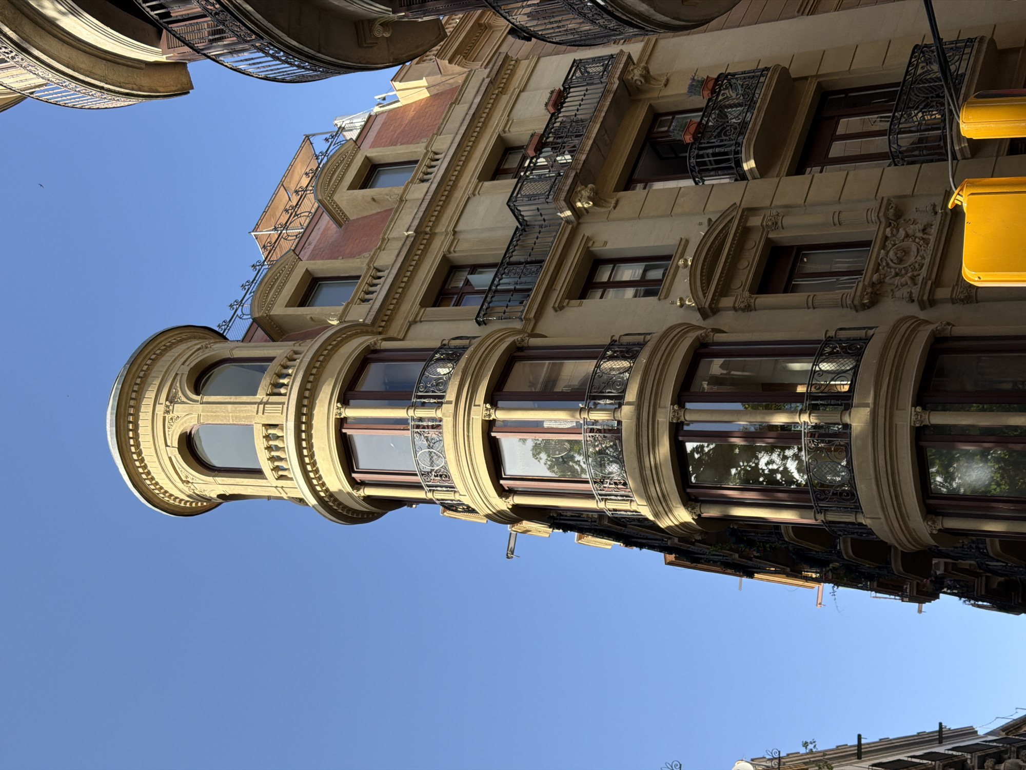 The image shows a sunlit building with a curved facade and a rounded turret on a clear blue sky. The building is made of light-colored stone and features several balconies with ornate black iron railings. The windows are tall and framed with dark wood. The architectural style appears to be Beaux-Arts or a similar late 19th or early 20th-century style. This building is located in the Eixample district of Barcelona, Spain. In the foreground, two yellow traffic lights are partially visible. The scene is devoid of people and focuses solely on the architecture and the sky.