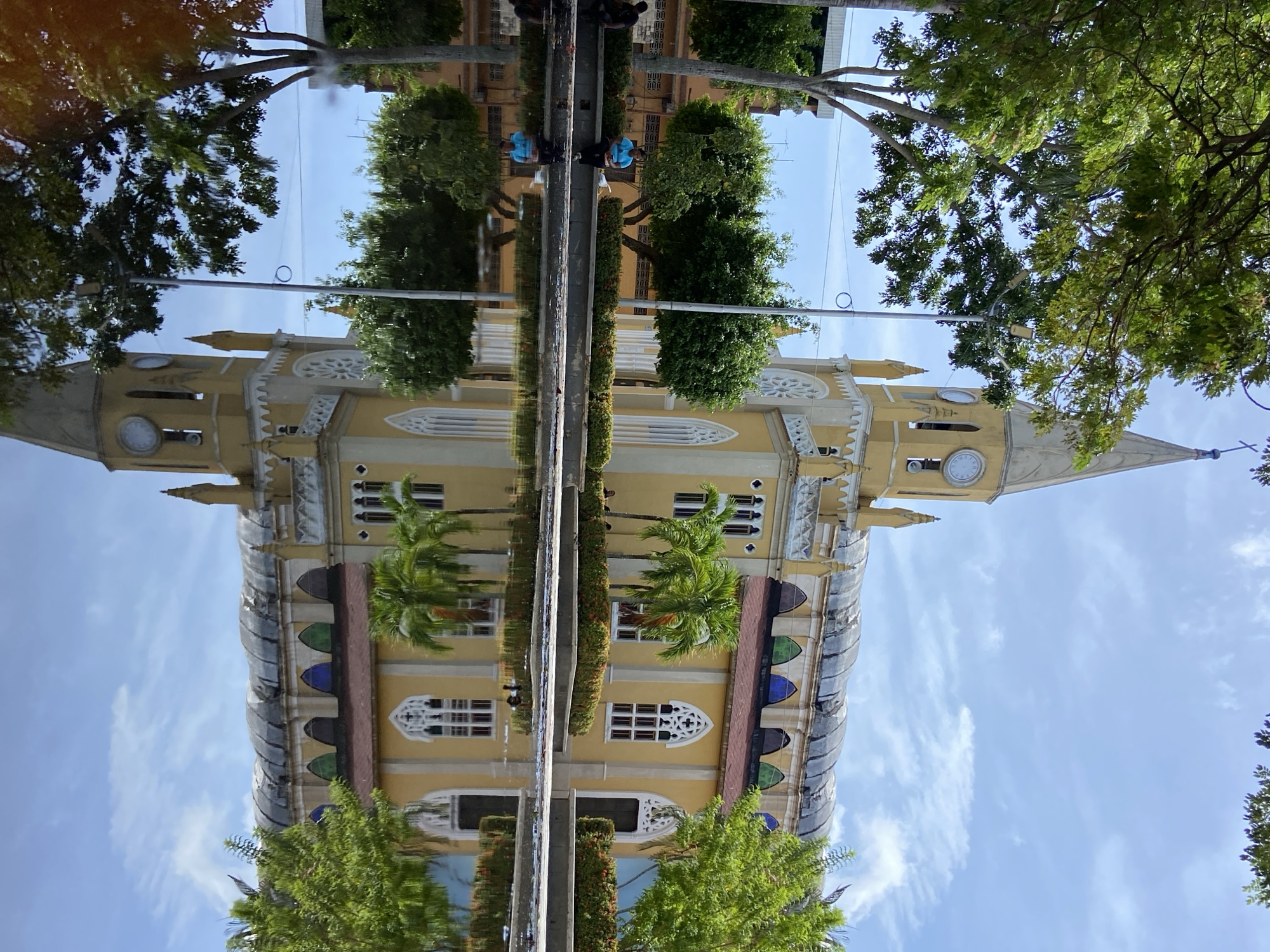 The image depicts a yellow church with a tall, pointed steeple and a clock face, possibly the Iglesia Nuestra Señora de la Soledad in San José de Barlovento, Venezuela. The church is reflected in a shallow body of water in the foreground, which also shows reflections of trees and the sky. There are trees and bushes bordering the church and the water. One person is visible sitting on the edge of the water, facing away from the camera, wearing a blue shirt. Another person is partially visible sitting on a ledge to the left. The sky is blue with scattered white clouds. The overall scene suggests an outdoor public space on a bright, possibly sunny day.