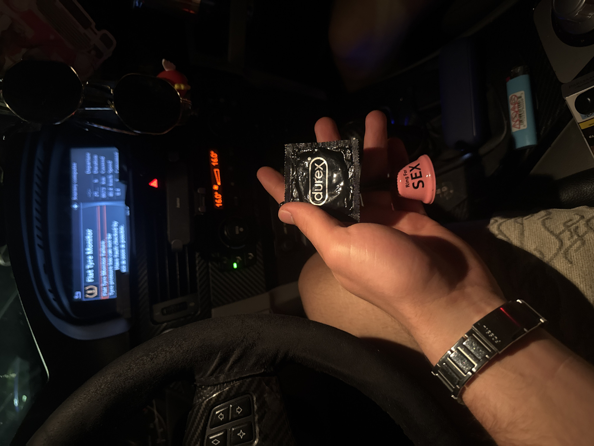 This media file captures a close-up perspective inside a car at night or in a dimly lit environment, likely in Varna, Bulgaria. The primary subject is a person's hand, presumably male, holding two items: a black Durex condom wrapper and a pink novelty bell with "Ring For SEX" written on it. The hand wears a silver-toned watch with a metal band, revealing the brand "FOSSIL" on its clasp.
The scene is set within the car's interior, featuring various elements of the dashboard and console. Visible details include a black steering wheel with carbon fiber accents and control buttons, a central display screen showing information from the "Journey Computer" and "Flat Tyre Monitor." The screen indicates a "Flat tyre monitor failure" and that "Tyre pressure has not been checked." Temperature readings of "16.0°" are displayed on the dashboard. Other objects scattered around the car include a pair of round sunglasses, a small red character figurine, a blue lighter with a warning label, and a blue box-like object. The overall ambiance is dark, with illuminated screens and light reflecting off various surfaces. The activity depicted is the holding and presentation of the condom and bell, suggesting a lighthearted or anticipatory moment.